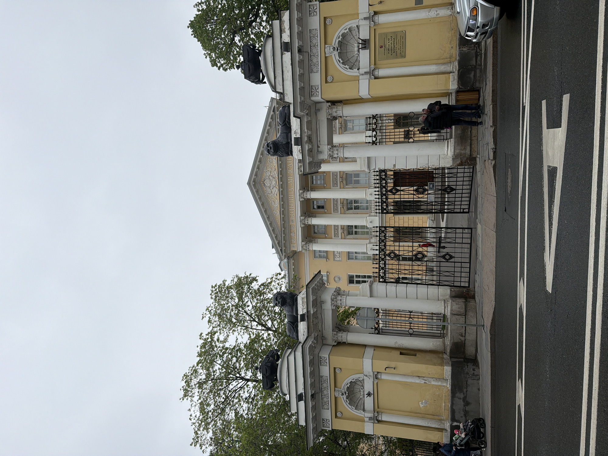 The image shows the entrance to the City Clinical Hospital No. 1 named after N.I. Pirogov in Moscow, Russia. The entrance is framed by yellow neoclassical structures adorned with lion statues. White columns support a facade. A black wrought-iron gate is visible in front of the building. Two people stand near the entrance, and a stroller is visible near the bottom of the image. A road with white lane markings is in the foreground. The sky is overcast.