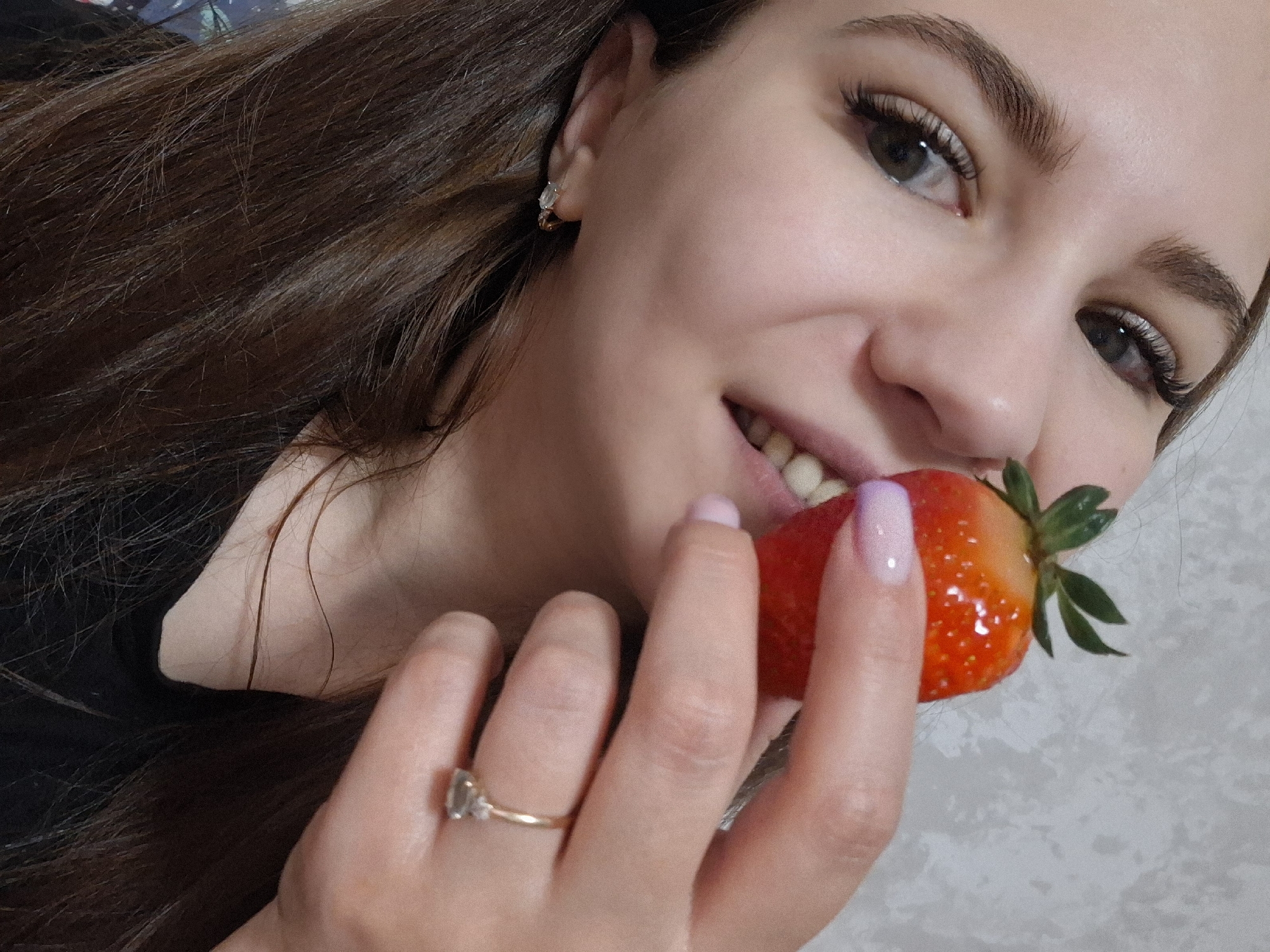 A young woman is holding a strawberry between her fingers and her mouth, as if she is about to eat it. She is smiling at the camera. She has brown hair, light skin, and wears a gold ring on her left hand. She has a small earring on the top of her right ear. The background is blurred and indistinct. There is no discernible information to determine the location is Chaikovsky, Russia.
