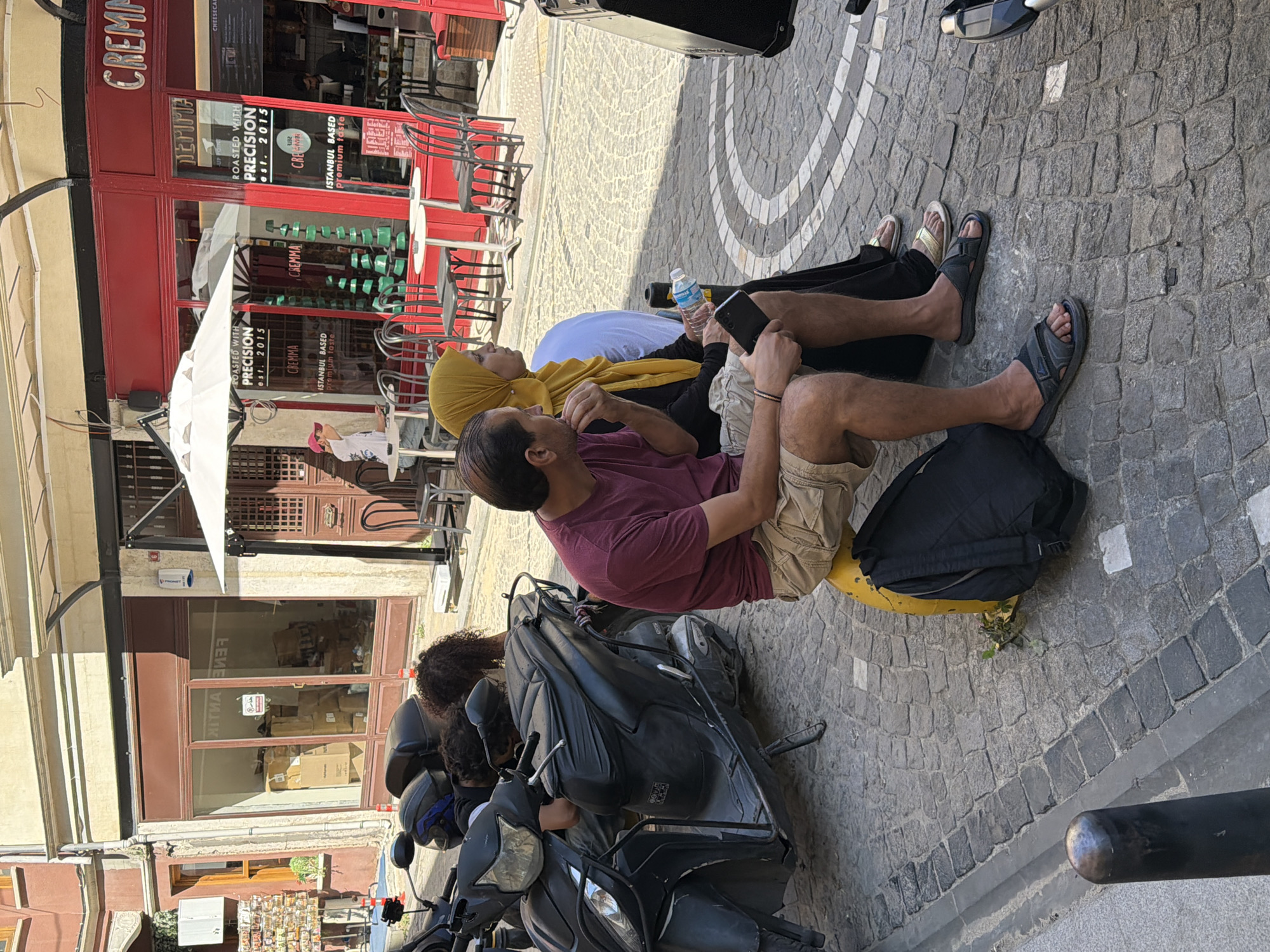 The image depicts a street scene in Istanbul, Turkey, with a cobblestone street and buildings lining the side. In the foreground, a man is sitting on a yellow cylindrical object, wearing a maroon t-shirt and khaki shorts. He is holding a smartphone. To his right, a woman wearing a yellow hijab and a black dress is seated, holding a bottle of water. They are facing away from the camera. A black scooter is parked to the left, with its front wheel visible. Behind the couple, a building with a red facade has storefronts with signage. One sign displays "CREMMA" and another reads "ROASTED WITH PRECISION est. 2015 ISTANBUL BASED premium taste." Outdoor seating with tables and chairs is visible in front of the storefronts. The overall atmosphere suggests a casual outdoor setting in a commercial area of the city.