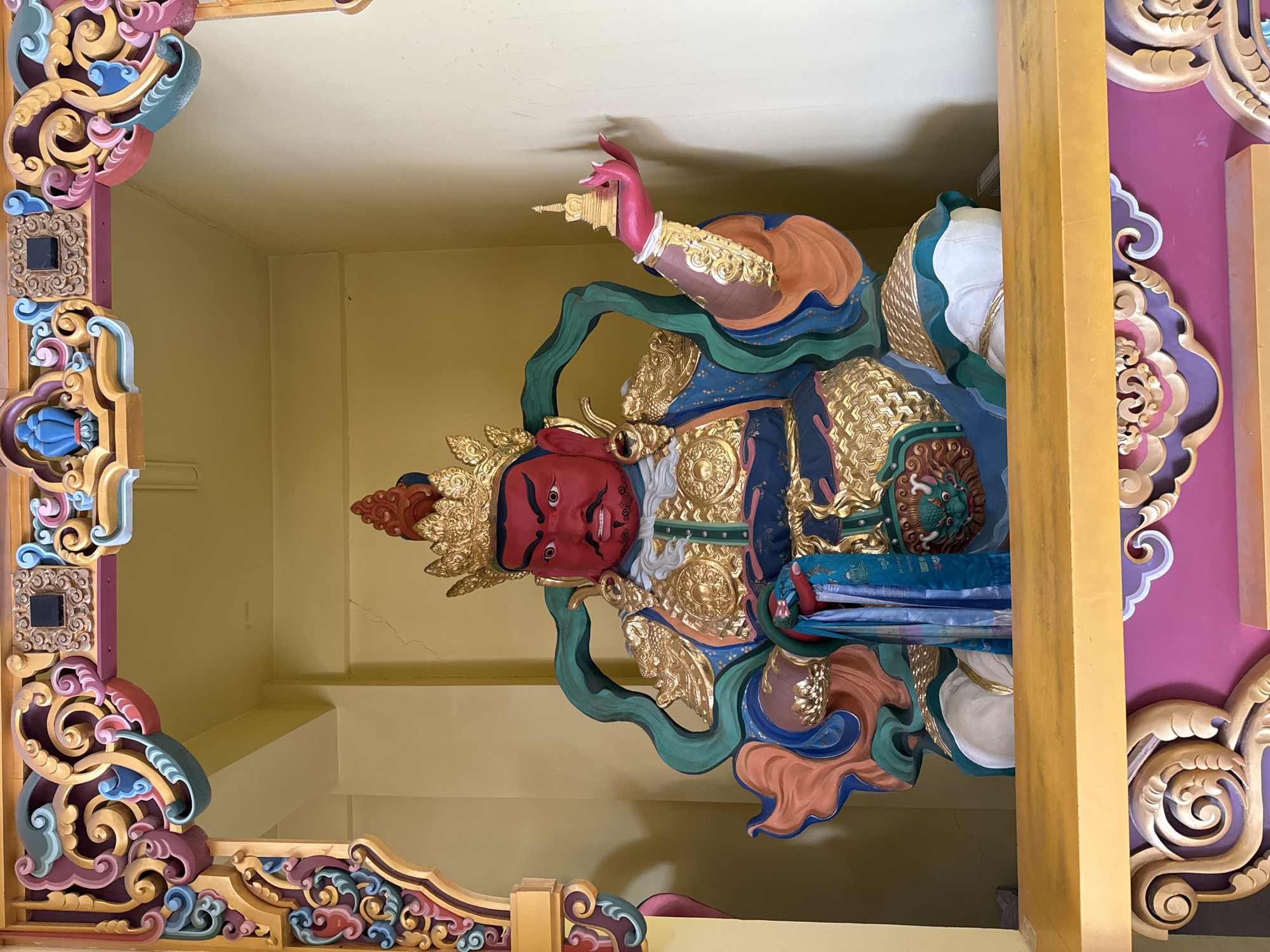 The image depicts a vibrant and detailed statue of a deity, adorned with gold and various colors, likely found within a temple or religious structure. The deity has a red face with a mustache and beard, wearing a golden crown. The body is adorned with ornate golden details over a blue and white robe. The statue is framed by elaborately carved and painted wooden architectural details, including a corner structure with colorful swirls and floral patterns, and a purple panel with cloud-like carvings. The location is indicated as being in Ulaanbaatar, Mongolia, suggesting the statue is likely part of a Buddhist temple or monastery within the city. The statue is positioned near the ceiling, further indicating it is part of a temple's interior architecture.
