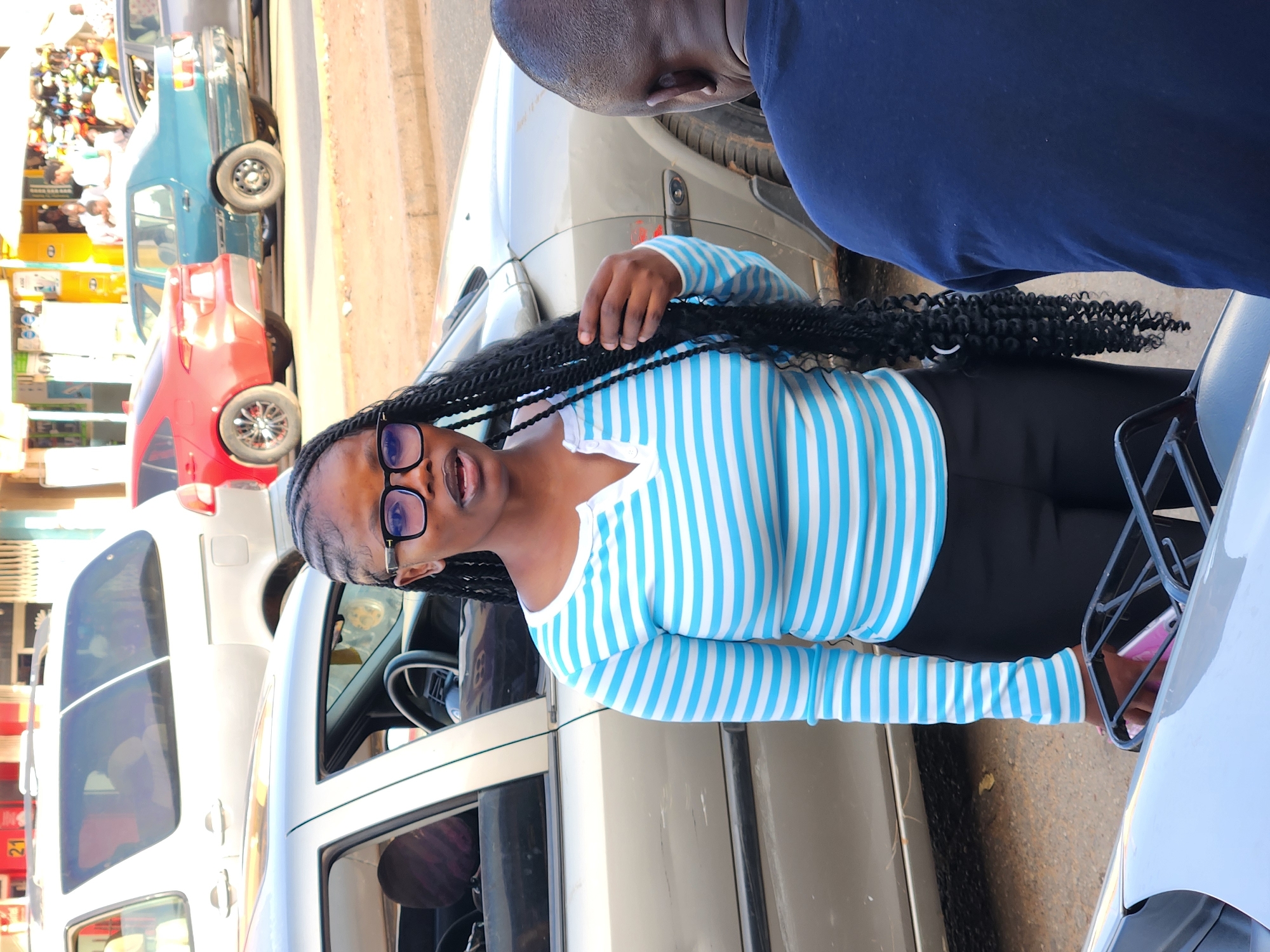 The image shows a woman in a blue and white striped shirt and black pants standing between two cars. She has long, dark braids and is wearing glasses. Her left hand is holding her hair. To her right is a black basket-like object with a pink cellphone inside. To the left of the image, a row of cars is visible, including a green, a red, and a white vehicle. Shops are seen on the very left. The person standing at the top is wearing a dark blue t-shirt. The city and location are unknown.