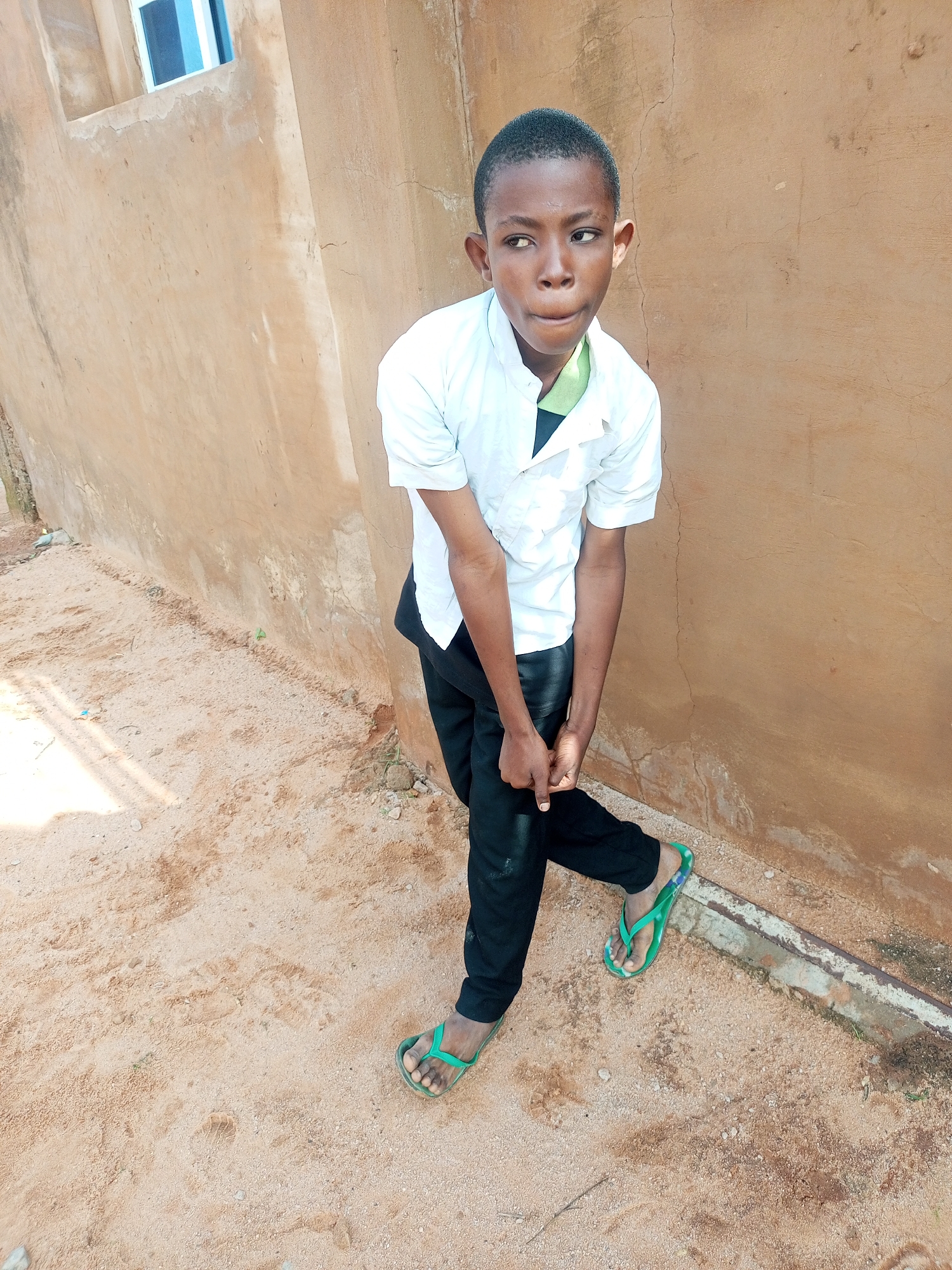 A young boy stands next to a tan, stucco wall in what appears to be an outdoor, urban environment. The boy is wearing a white button-down shirt and black pants, with green flip-flops on his feet. He is looking to his left, with his hands clasped in front of him. The ground is covered in light brown sand and small pebbles. A sliver of a blue and white window can be seen on the wall behind the boy.