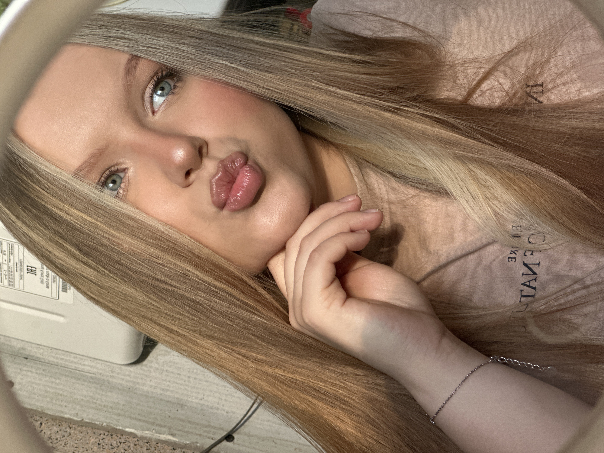 A young woman with blonde hair is posing for a selfie. She has light blue eyes, fair skin, and is wearing makeup. She is pouting her lips and resting her chin on her hand, which is adorned with a thin silver bracelet. She is wearing a light pink t-shirt. A ring light is partially visible around her. The background appears to be an indoor setting with a white appliance with labels, and part of a gray surface. The location in the city of Chaikovsky, Russia, is not determined from the image.
