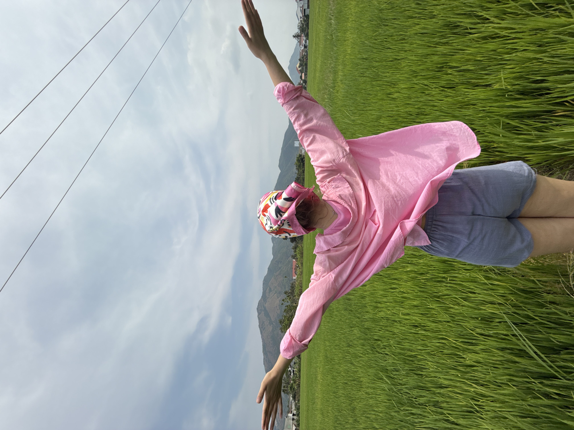 A person stands amidst a lush, verdant rice field, with arms outstretched wide, conveying a sense of freedom and embrace of the natural surroundings. The individual wears a bright pink long-sleeved shirt, light blue or grey shorts, and a colorful patterned bandana or head covering featuring shades of pink, red, yellow, and white.
The scene unfolds under a partially cloudy sky, with several thin utility wires traversing the upper left portion of the frame. In the background, rolling green mountains rise majestically, and at their base, scattered clusters of residential buildings or a village are visible, nestled among trees. The foreground and midground are dominated by the vibrant green of the rice paddies, stretching towards the distant mountains.
The time of day appears to be daytime, possibly late morning or early afternoon, characterized by soft, natural light. The weather is clear and pleasant, with white clouds scattered across the sky. The overall atmosphere evokes tranquility and joy, highlighting a connection with nature in a serene, rural landscape. The visual cues, including the rice fields and mountains, are consistent with the provided location context of Diên Khánh, Vietnam. There is no visible text in the image.