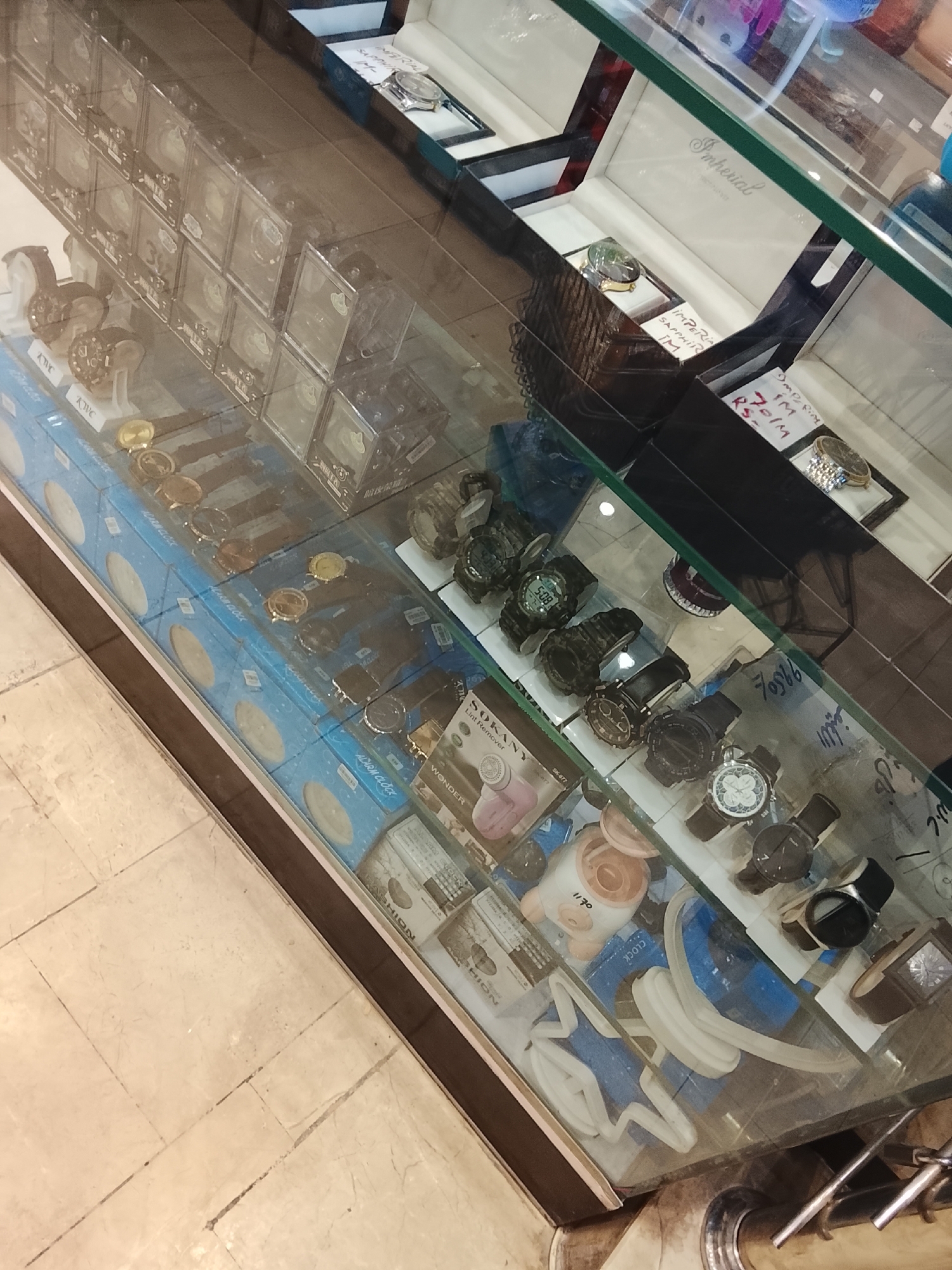 A display case in Gulgasht Colony, Pakistan, is filled with various items, primarily watches and watch-related products. The items are arranged on different levels within the glass case. On the top level are watches in display boxes. Below this, there are watches packaged in blue boxes labeled "ALARM CLOCK." Another level contains a collection of watches with various designs. There are also lint removers by "SOKANY" and other accessories, along with displays that appear to be watch-related. The lighting is a bit dim, and there are price tags on some of the items. The case is made of glass and is possibly part of a retail setting.