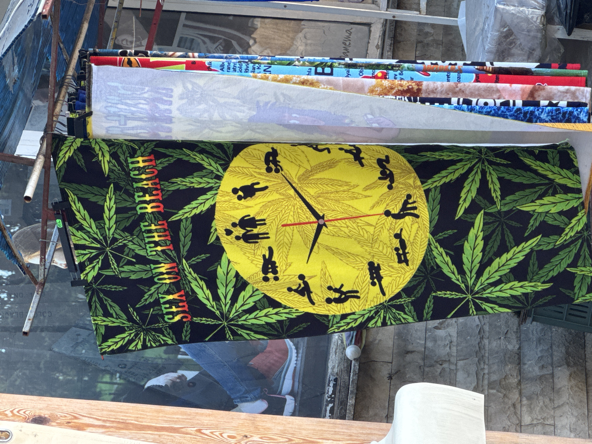 The image shows a street market scene in Tsarevo, Bulgaria. In the foreground, a black towel with green cannabis leaves and the text "SEX ON THE BEACH" in red and yellow letters is displayed. In the center of the towel is a yellow clock face with black silhouettes of people in various sexual positions representing the hours. To the right of the towel are other textiles hanging, with colorful patterns and text, including what appears to be a towel with the word "Panema" written on it, possibly referencing the Copacabana beach in Rio de Janeiro. In the background, a person's legs wearing blue jeans and red sneakers are visible, suggesting they are a vendor or shopper. The market appears to be set up outdoors, with a rustic metal structure overhead supporting the hanging items. The ground is a stone surface, and a green plastic crate is visible. The overall impression is a vibrant, somewhat provocative display typical of a seaside tourist town.