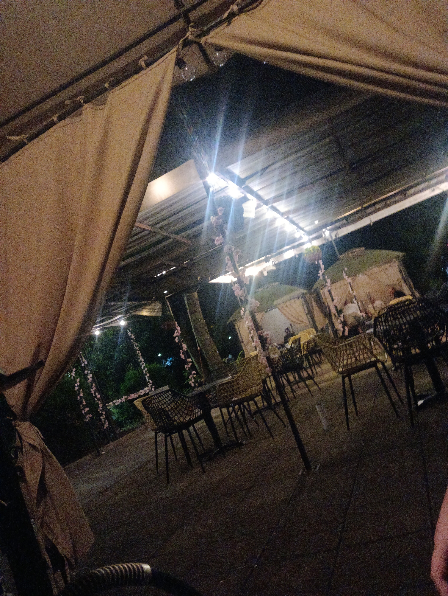 The image depicts an outdoor seating area at night, possibly a restaurant or cafe. Several tables and chairs are visible, along with covered seating areas or cabanas. String lights illuminate the scene, casting bright flares. Decorative floral garlands hang in some areas. The ground appears to be paved. The overall impression is that of a dimly lit, atmospheric outdoor dining space. The user mentioned Burgas, Bulgaria, but there are no specific visual cues in the image to confirm this location.