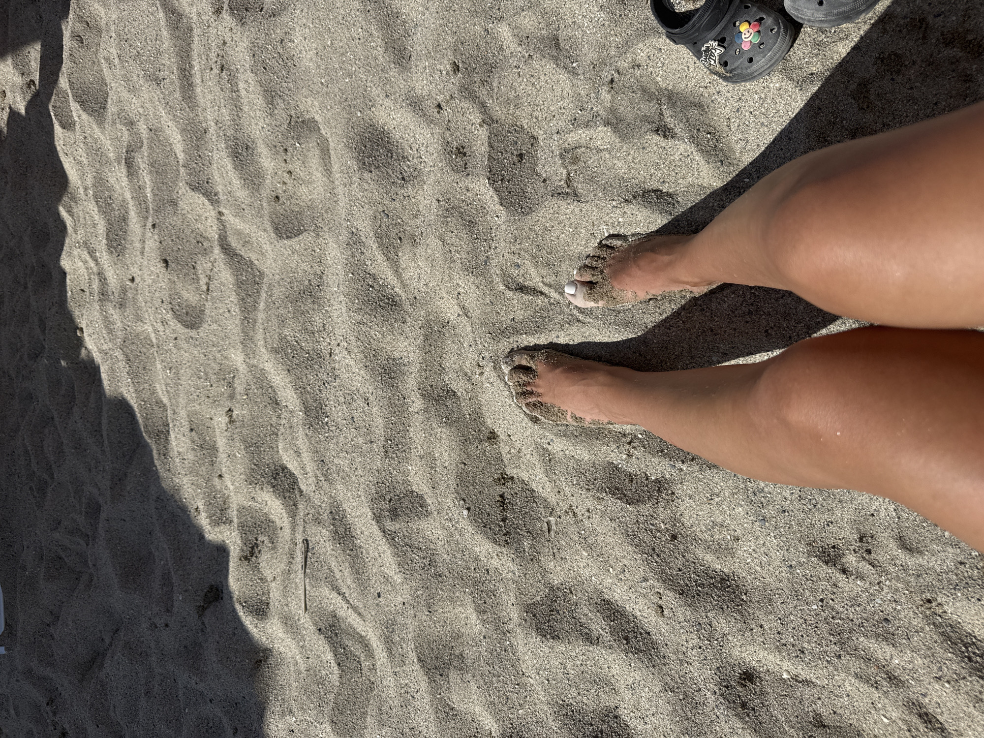 The image depicts a close-up, top-down view of a person's legs and feet in the sand, likely on a beach in Finale Ligure, Italy. The sand is a light gray color with visible ripples and texture, suggesting it is a natural beach. The person's legs are tanned, and their feet are partially covered in sand. One foot has white nail polish on the toenails. To the right of the feet, a black Croc shoe with a colorful charm is visible. A shadow, likely from an umbrella or awning, casts a diagonal line across the scene. The lighting suggests it is a sunny day.