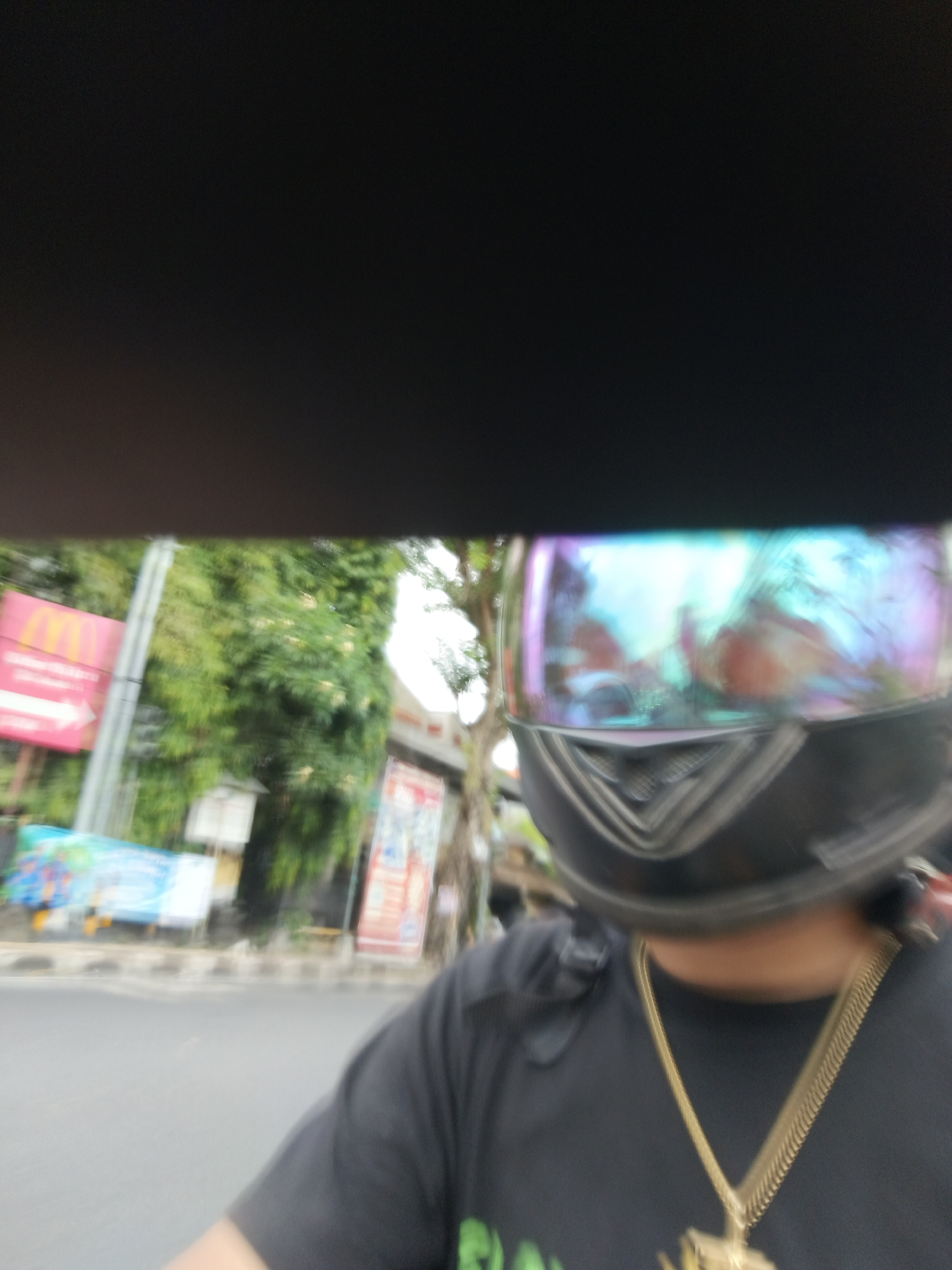 In the foreground, a person wearing a black t-shirt and a black helmet with a iridescent visor is riding a motorcycle. A gold chain with a pendant is visible around the person's neck. The background shows a street scene in Denpasar, Indonesia, with blurred trees, buildings, and street signs, including what appears to be a McDonald's sign. The road is visible, with traffic markings and possibly other vehicles in the distance. The image is slightly motion-blurred, suggesting movement.