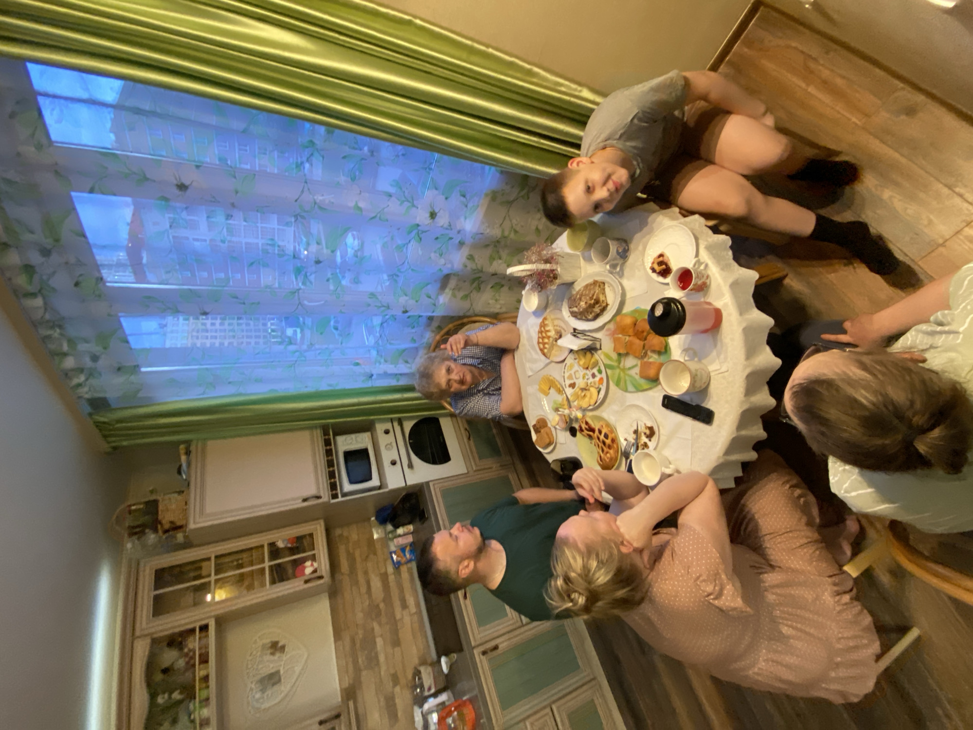 A slightly rotated medium shot captures a family meal in what appears to be an apartment in Yekaterinburg, Russia. Four adults and one child are seated around a round, white-clothed table laden with food, including pastries, fruit, and beverages in cups and containers. An older woman is seated to the left of the frame, wearing a blue-and-white checkered shirt, and a younger man in a green shirt is seated to her right. Another woman wearing a pink polka-dot dress sits adjacent to the man. Opposite her is another woman with short brown hair wearing a light green shirt. A young boy with short brown hair and a gray shirt is seated on the floor to the upper right of the table.
The setting is a kitchen with light-colored cabinets and appliances, including a microwave and oven. Light filters through a window with patterned blinds. The floor is wood.