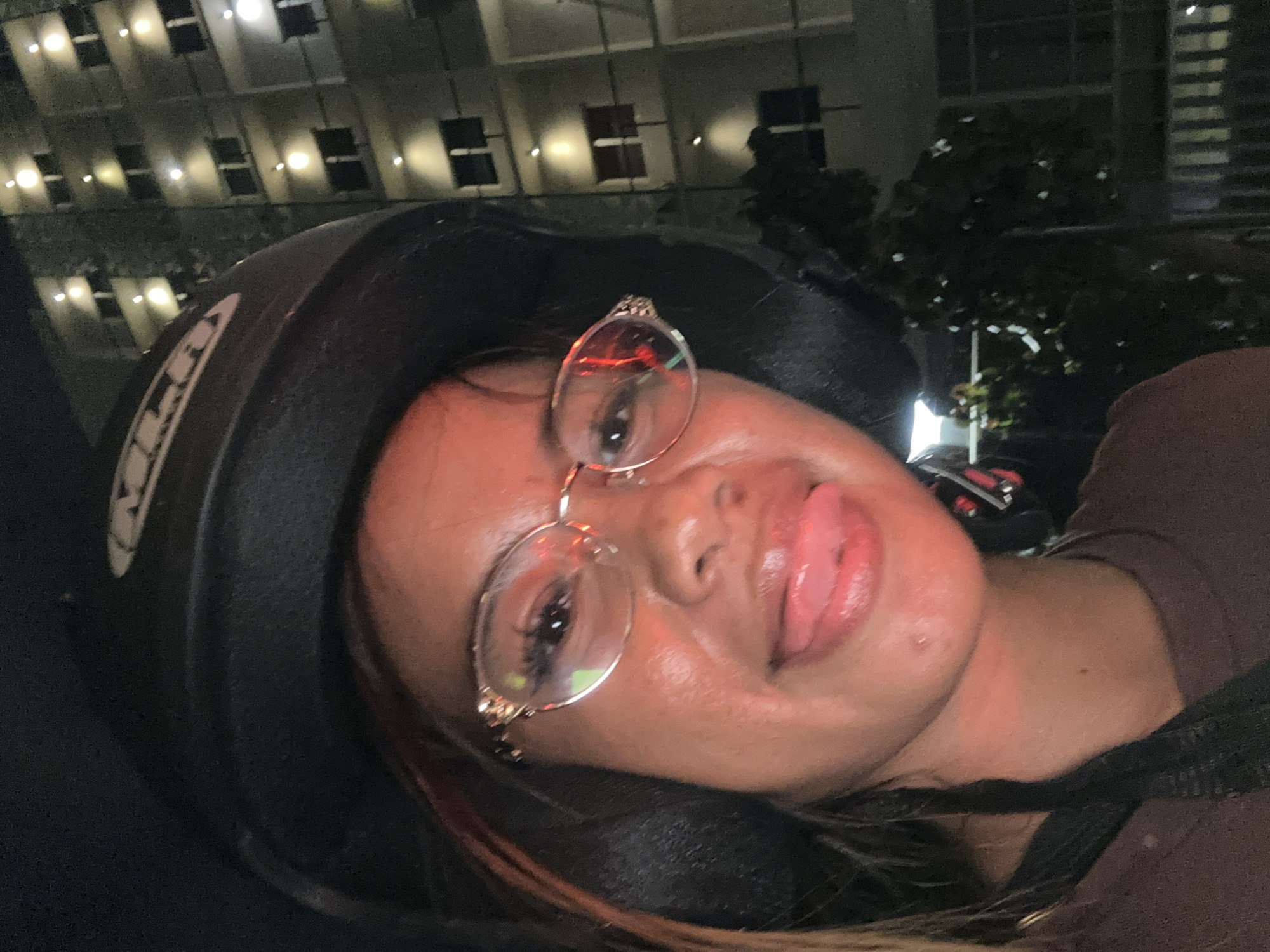 A close-up, angled shot features a person wearing a motorcycle helmet and glasses, sticking out their tongue. Buildings with illuminated windows are visible in the background, suggesting an urban environment. The location appears to be Central Jakarta, Indonesia.