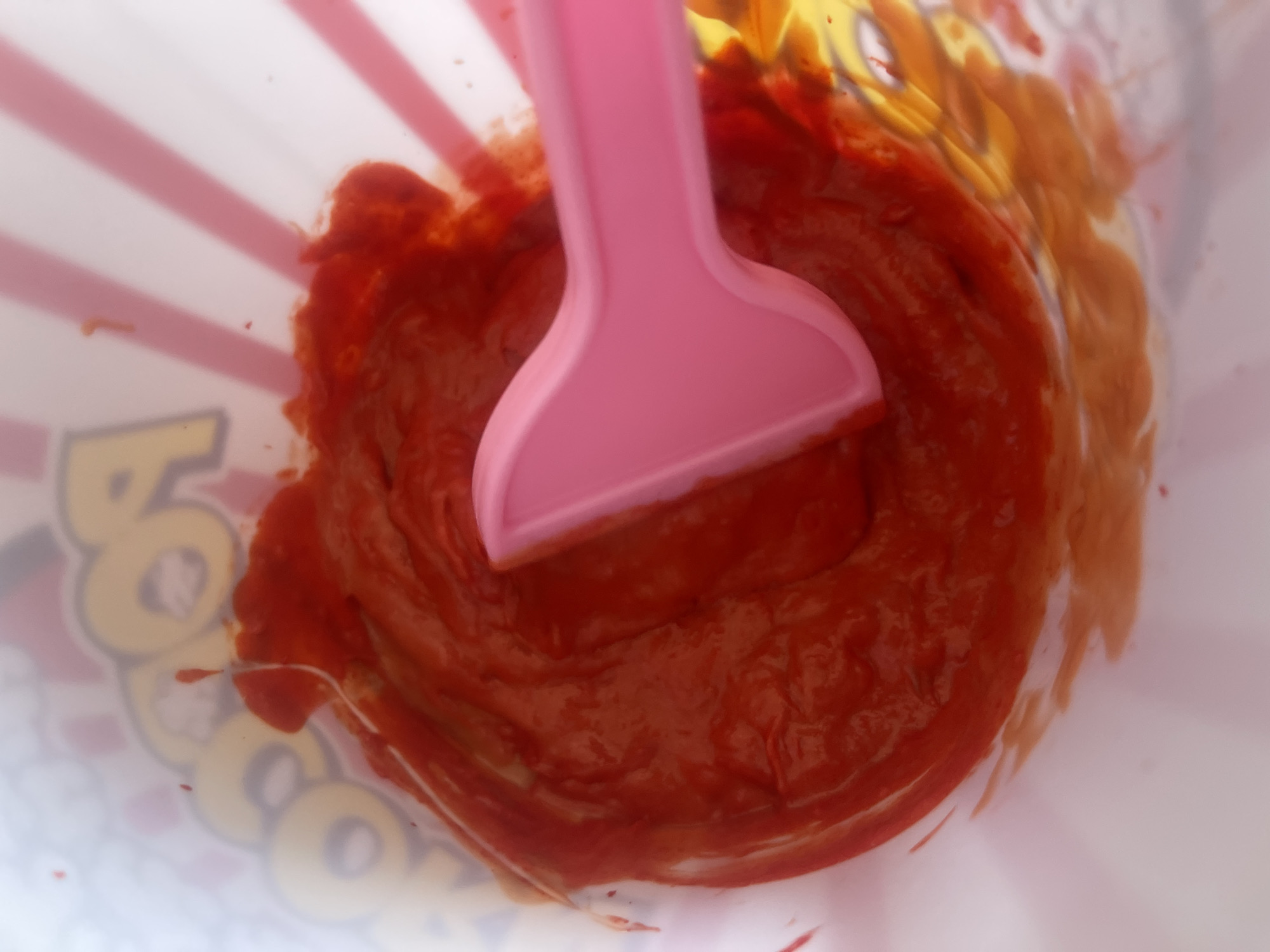 The image shows a close-up view of a pink plastic spoon submerged in a vibrant red substance, possibly a sauce or dip, inside a popcorn container. The container has a white background with red stripes and yellow lettering that reads "POP" which is partially visible. The red substance appears thick and has streaks and swirls from being mixed. The overall impression is of a processed food item. There is no indication of location in Varna, Bulgaria or any people present.