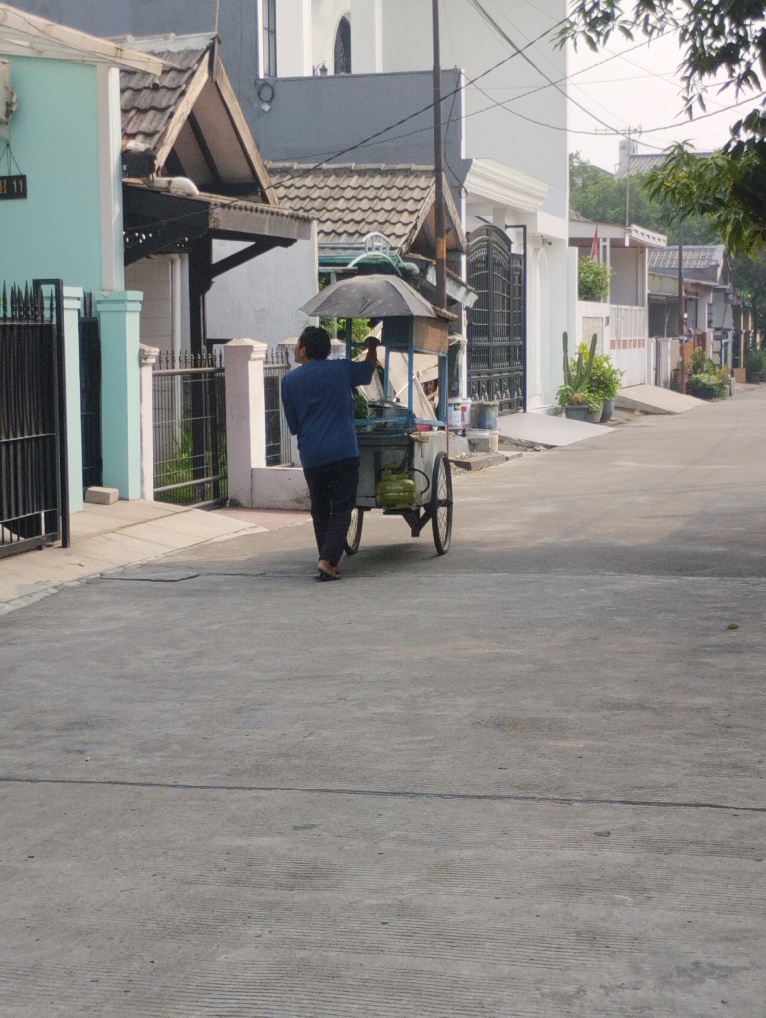 A man in a blue shirt and dark pants pushes a food cart down a paved street in Bekasi, Indonesia. The food cart has an umbrella and a visible gas tank. The street is lined with houses and fences. The weather appears to be sunny with some trees and foliage visible in the background. The scene is captured from a slightly elevated perspective, looking down at the street.