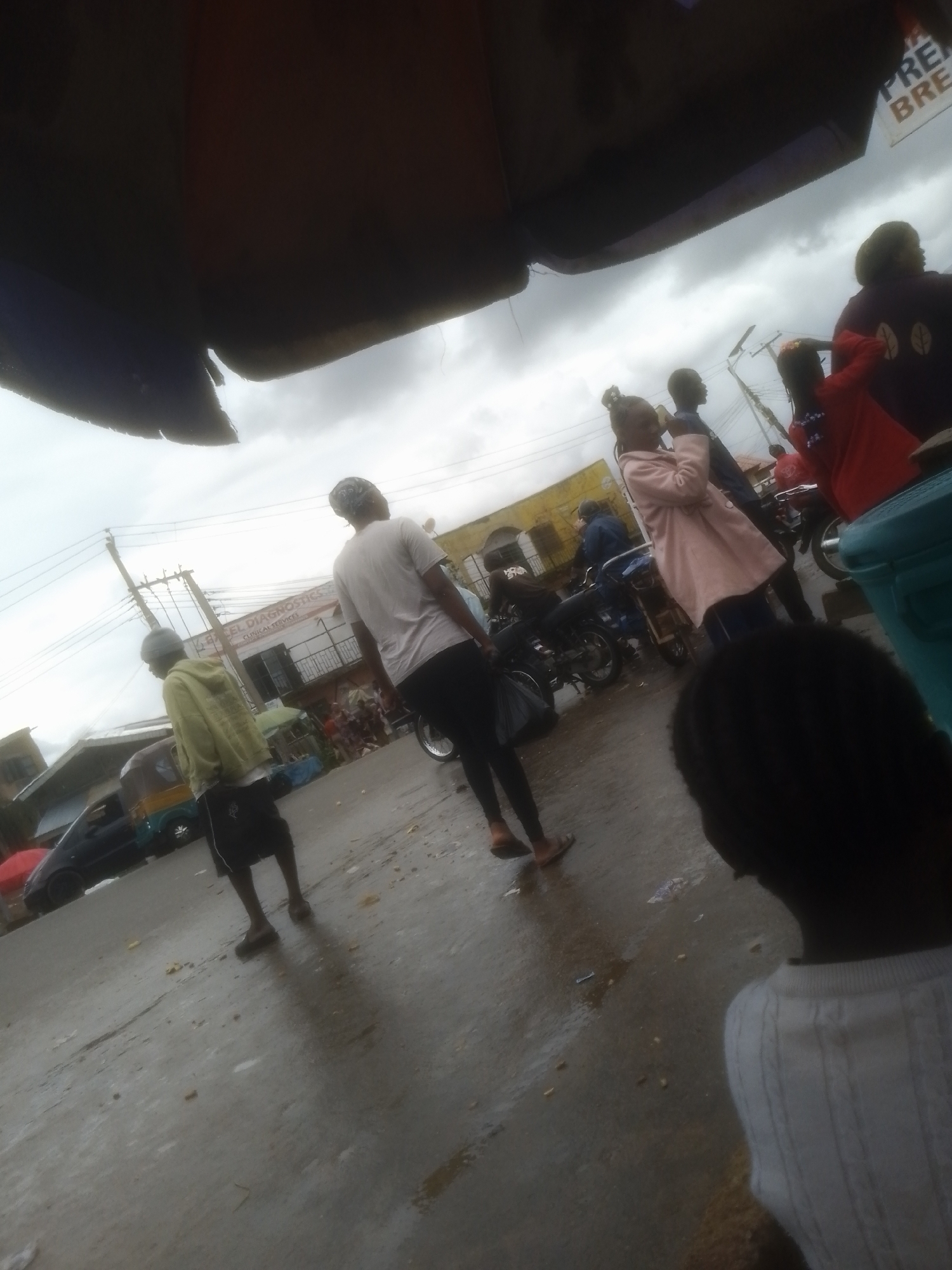 The image depicts an outdoor scene, likely a street market or busy thoroughfare, under a cloudy sky. The environment appears somewhat damp, suggesting recent rain. In the foreground, a person's back is visible, wearing a white knitted sweater. Further back, a group of individuals are present. One person, seen from behind and in profile, is wearing a light-colored hooded jacket and dark shorts, with a grey beanie. Another individual, also viewed from behind, is wearing a white t-shirt and dark pants. A woman in a light pink coat is walking, and behind her, a person in a red outfit. Several motorcycles are parked and in use, with one being ridden by a person in a blue shirt. In the background, buildings and a sign for "BARCEL DIAGNOSTICS CLINICAL SERVICES" are visible. A dark-colored car and a three-wheeled vehicle are also present. The overall atmosphere suggests a moment captured in a bustling urban environment in Gwong, Nigeria.