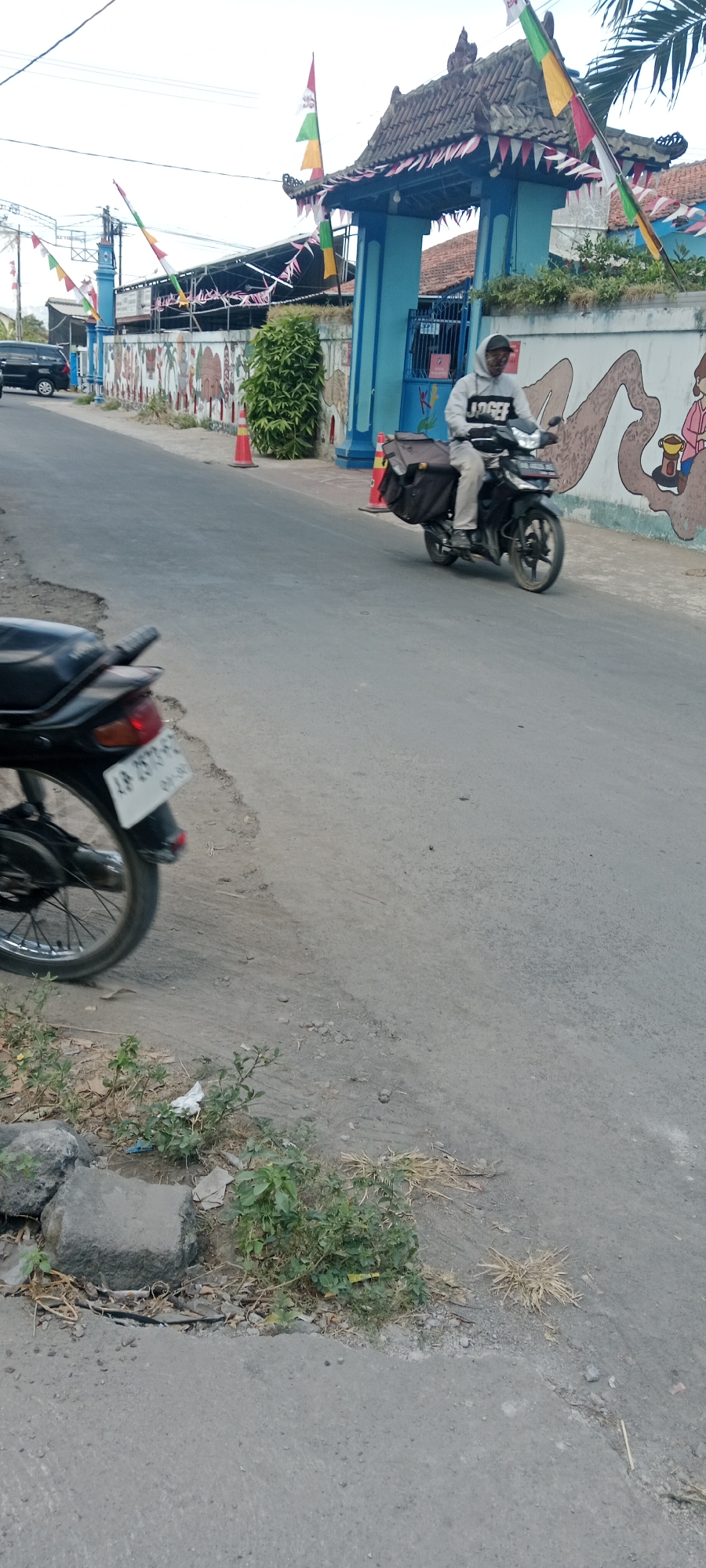 In Purwomartani, Indonesia, a man wearing a grey hooded sweatshirt rides a black motorcycle with a large black cargo bag strapped to the rear. The motorcycle is on a paved road in front of a building with a blue gate and a white wall decorated with colorful murals. The building has a traditional Indonesian roof structure adorned with small flags. Red and orange traffic cones are placed along the roadside. In the foreground, the rear wheel of another black motorcycle is visible, with a license plate that reads "AP 2573 FC". The environment is a street with a worn asphalt surface, bordered by a curb and patches of dry vegetation and rocks. The sky is overcast.