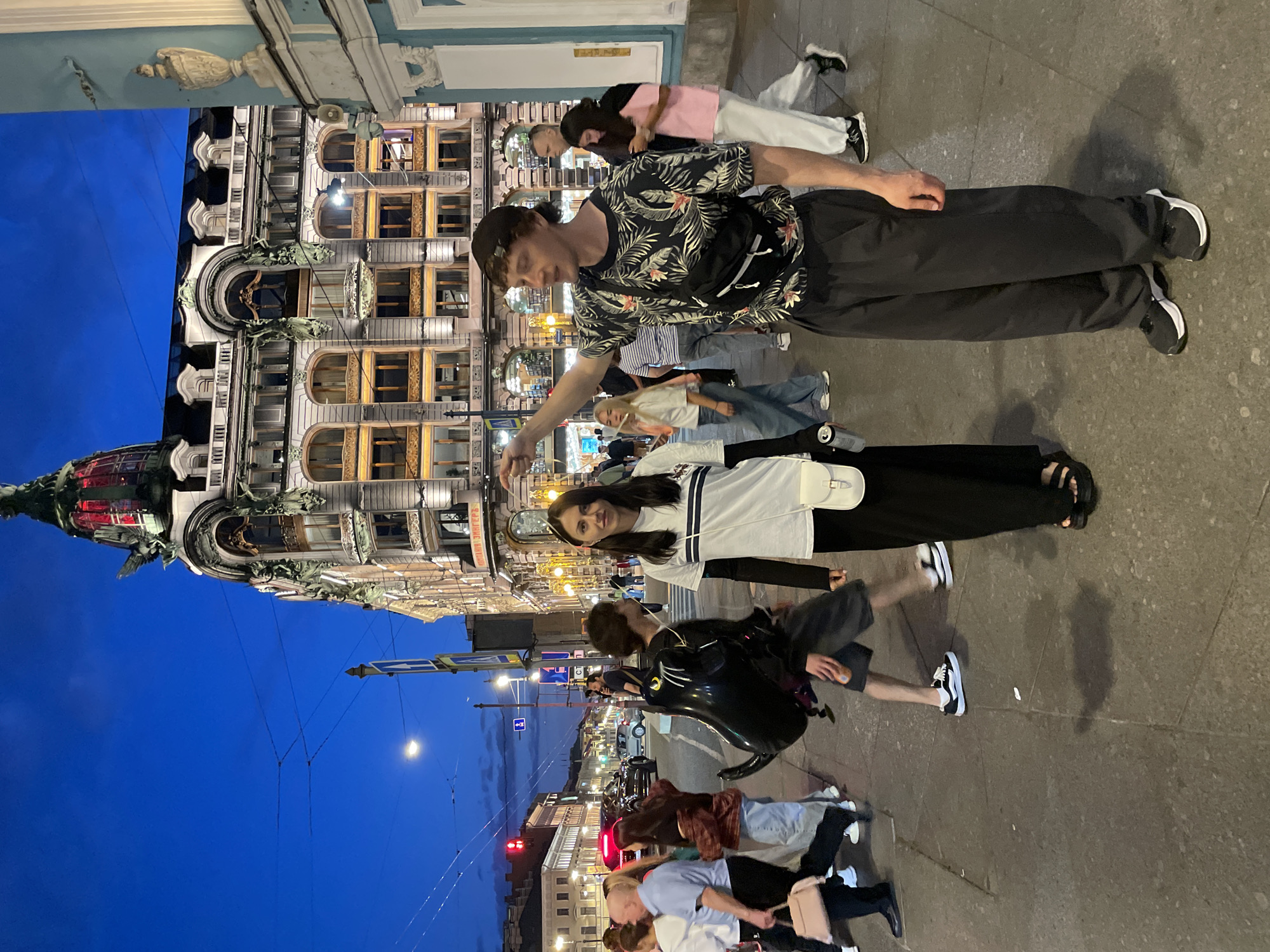 This image depicts an early evening urban street scene in St. Petersburg, Russia, specifically focusing on the area around the historic House of Singer (Dom Knigi) building. The sky is a deep twilight blue with faint cloud formations, and artificial lights illuminate the street and buildings. In the foreground, two individuals stand on a paved sidewalk, facing the camera. On the right, a man with a dark baseball cap worn backward is seen from the waist up. He wears a black short-sleeved shirt with a green leaf and pink/orange floral pattern, dark loose-fitting trousers, and black athletic shoes with white soles. A black fanny pack is slung across his chest. His right arm is extended, and his index finger points towards the left side of the frame. To his left, a woman stands facing the camera with a neutral expression. She wears a white short-sleeved t-shirt with two thin horizontal dark stripes over a long-sleeved dark underlayer, long black trousers, and black sandals. A small white shoulder bag hangs from her left shoulder, and she holds a silver can in her left hand. The mid-ground features several pedestrians. To the left of the central woman, a person walks right-to-left, carrying a large, inflated black balloon shaped like a panther or cat. They wear dark shorts and sneakers. Further back, various other individuals walk in different directions, some appearing blurred due to motion. A man in a light blue collared shirt and dark pants walks leftward on the far left, carrying a light pink handbag. A woman in a pink top and light pants walks away from the camera on the right side of the frame. Vehicles, including cars, are visible on the street in the distance. The background is dominated by the ornate House of Singer building on the right. This multi-story structure features classical architectural elements, including large arched windows, decorative green-patinated sculptures, and exterior illumination. The building's dome has a red-lit section and is topped with a large sculpture of an eagle. A sign on the building's facade reads "КОМПАНІЯ ЗИНГЕРЪ". To the right of the Singer building, a lighter blue/grey building with a white decorative urn is partially visible. On the left side of the street, other multi-story buildings line the road. Overhead electrical wires crisscross the sky. A blue and white street sign is visible on a pole near the center-left, and a streetlight illuminates the