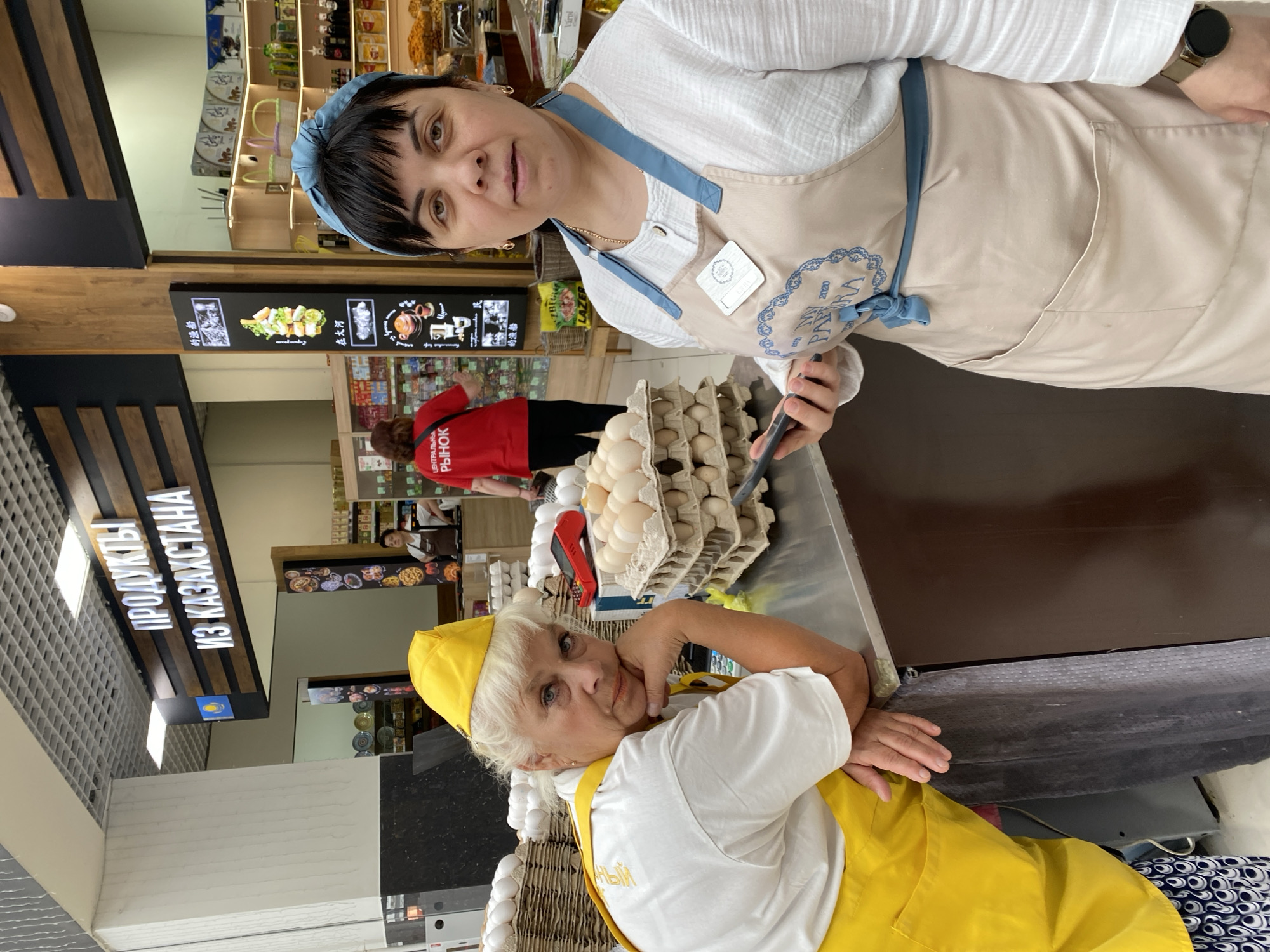 Two women are posing for a photograph in a store that sells food products from Kazakhstan, likely in Novaya Usman', Russia. The woman on the left, with blonde hair, wears a yellow hat and apron over a white shirt. She is leaning on her elbows on a counter, looking at the camera with a slightly stern expression. In front of her are several stacks of egg cartons filled with eggs. The woman on the right, with dark hair, wears a blue headband and a beige apron over a white shirt. She is holding a smartphone in her right hand and looking directly at the camera. Behind the women, shelves are stocked with various food items, and signage indicates "Продукты из Казахстана" (Products from Kazakhstan) and "Центральный Рынок" (Central Market). The environment appears to be a retail grocery store.