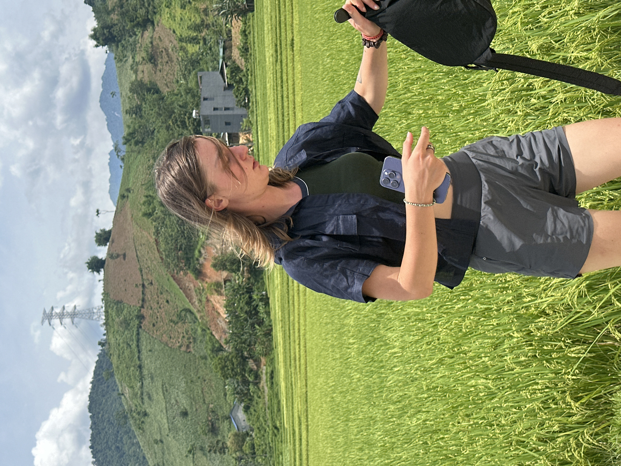 This image captures a person standing amidst vibrant green rice paddies in Duong Quy, Vietnam, under a bright, partly cloudy daytime sky. The subject, a female with blonde or light brown hair, is positioned slightly to the left of the frame, facing forward and looking slightly downwards. She is dressed in a dark blue short-sleeved button-up shirt over a dark green top, paired with dark grey shorts.
In her right hand, she holds the strap of a black bag. Her left hand holds a purple smartphone, identifiable by its distinct triple-lens camera design, typical of a newer iPhone model. She accessorizes with a red and dark-colored bracelet on her right wrist, a light-colored beaded bracelet on her left wrist, and a ring on her left hand's ring finger. Her expression is neutral, possibly thoughtful or squinting slightly due to the bright light.
The background features expansive, lush green rice fields that form terraces across rolling hills and mountains. These hills are covered in a mix of dense vegetation and some visible patches of reddish-brown earth. A modern, multi-story grey building is visible nestled among the distant greenery, and a tall metal electrical transmission tower stands on a far-off hillside. The scene is illuminated by clear daylight, suggesting it is late morning or early afternoon.