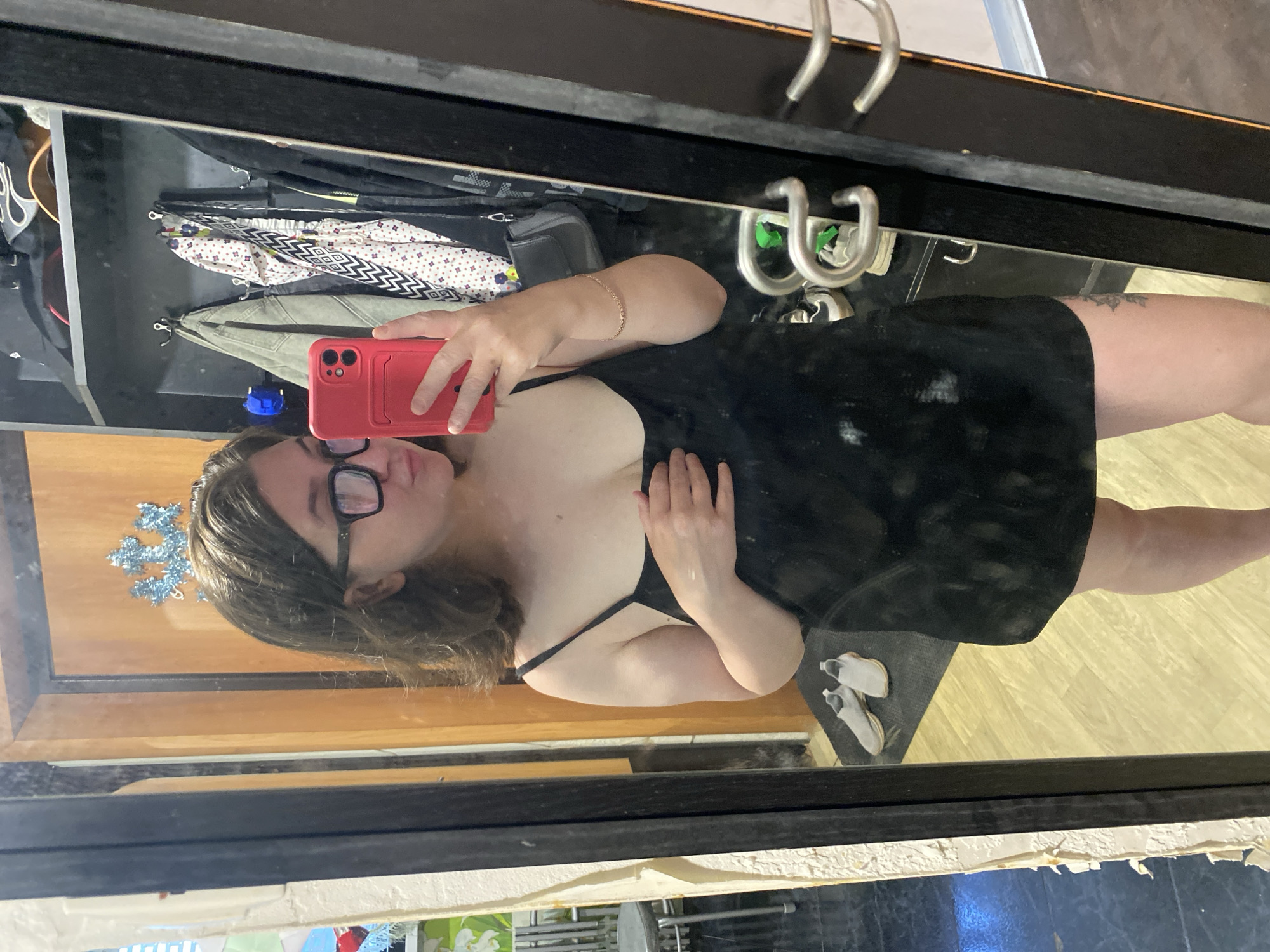 A woman with glasses and a black dress takes a mirror selfie with a red phone. In the background, a closet with clothes and hangers is visible. There is a wood floor in the background, and outside the window below the mirror, a plant is visible. The location is presumably in Chaikovsky, Russia.