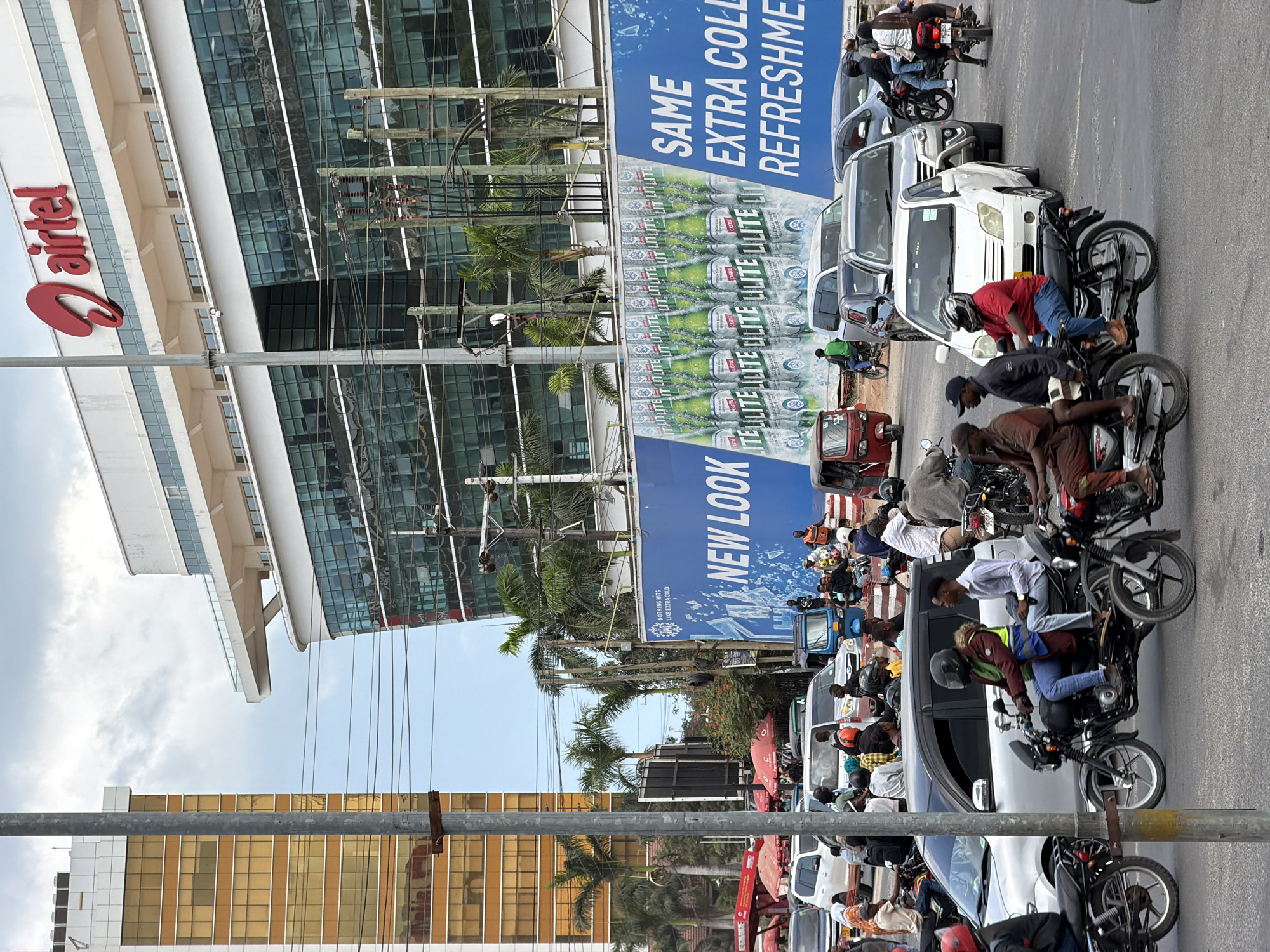 The image captures a bustling street scene in Dar es Salaam, Tanzania, featuring a prominent Airtel building in the background. The modern, glass-fronted Airtel building stands tall, adorned with the company's iconic red logo. In front of it, a large blue billboard advertises "NEW LOOK" and "SAME EXTRA COLD REFRESHMENT," showcasing bottles of Castle Lite beer.
The street is crowded with various modes of transportation. Several motorcycles, some carrying multiple passengers, navigate through the traffic. Cars, including a white SUV and a silver sedan, are also present. A few tuk-tuks, a common form of transport in Dar es Salaam, can be seen. The sidewalks are lined with people, some walking, while others are gathered, possibly waiting for transportation or engaging in conversations. Palm trees are scattered throughout the scene, adding a tropical touch to the urban environment. The sky is overcast, suggesting a cloudy day. The overall atmosphere is one of a busy city day in Dar es Salaam.
