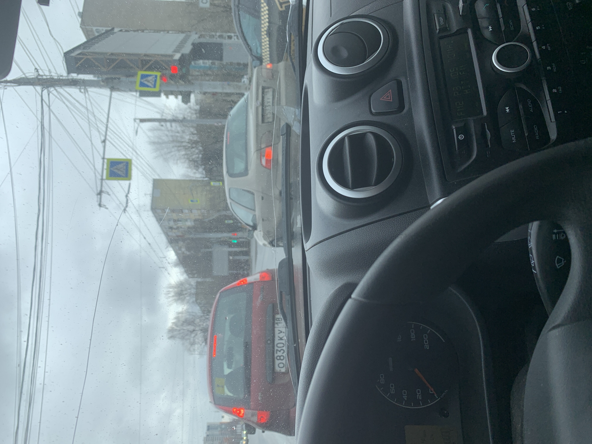 The image is taken from inside a car in Izhevsk, Russia, during what appears to be a cloudy or overcast day. The car is stopped in traffic. A red car in front has a license plate "O830KY 18," which is a Russian vehicle registration. Traffic signs for pedestrian crossings are visible in the distance. The car's dashboard and steering wheel are in the foreground, with the radio displaying a frequency.