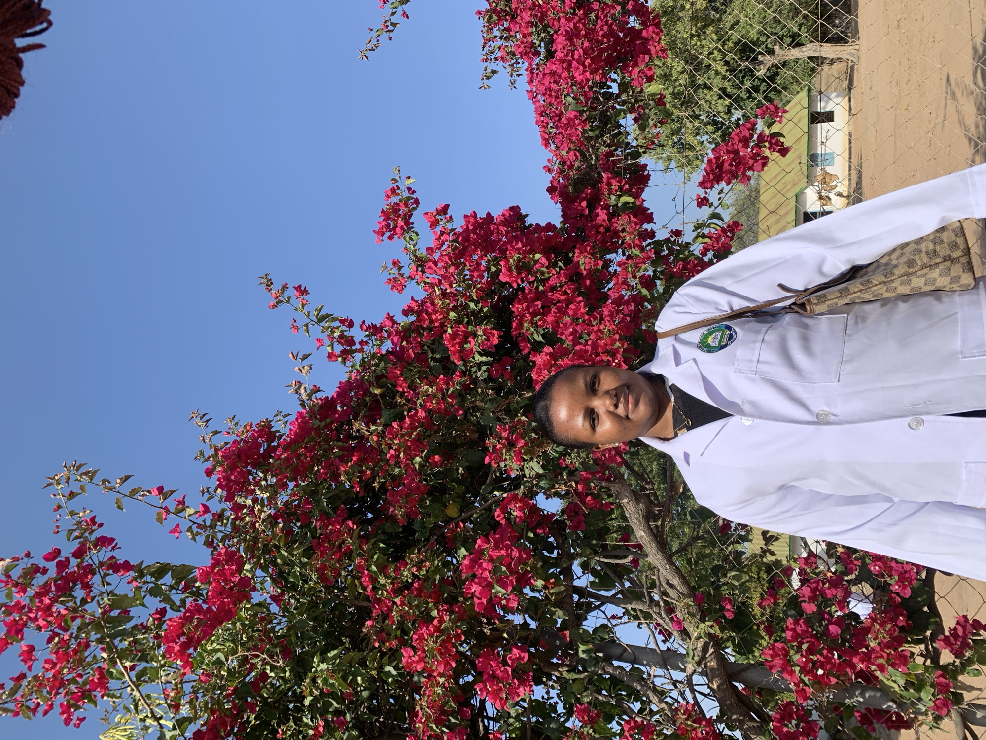 A medium shot shows a young Black woman wearing a white lab coat with a crest on the left breast, a black shirt underneath, and a necklace. She is standing outside in front of a vibrant bougainvillea tree with bright pink flowers and green leaves. The background includes a chain-link fence, a small, white building with a green roof, and a clear blue sky. The woman is looking directly at the camera with a slight smile. The overall impression is one of a bright, sunny day in a somewhat rural or developing area, possibly a clinic or educational institution, given the lab coat.