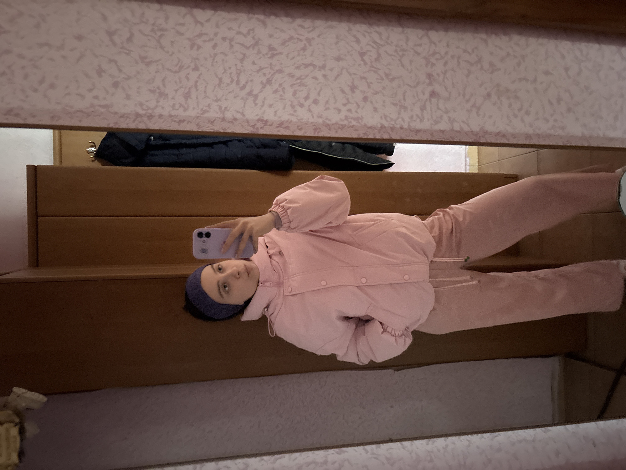 A person wearing a pink tracksuit and a blue headband is taking a selfie with a purple phone in front of a mirror. The background shows a wooden structure, a black jacket hanging above, and a patterned wall. The location is Прикамский, Russia.