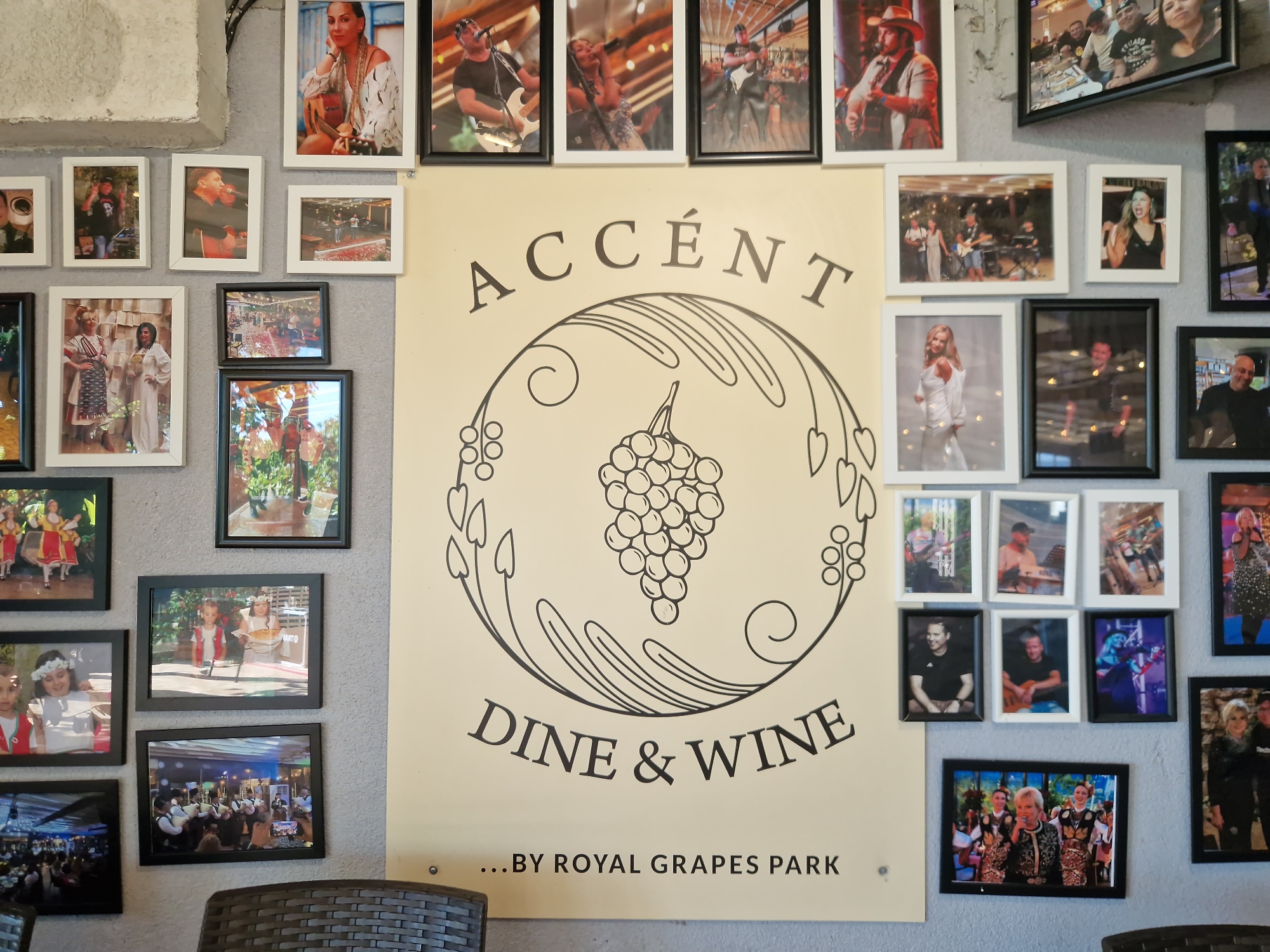 The image is a medium shot of a wall in a venue, likely a restaurant or a performance space, in Tankovo, Bulgaria. The wall is adorned with numerous framed photographs and a large poster. The poster, centrally located, features the text "ACCÉNT DINE & WINE ...BY ROYAL GRAPES PARK" in a stylized font, with a graphic of grapes and leaves. The photographs are arranged in a grid-like fashion around the poster. Many of the photos depict people, some of whom appear to be musicians performing, while others are candid shots of individuals or groups. The overall impression is of a decorated space that celebrates music and gatherings, possibly with a connection to a winery or vineyard given the poster's text. The lighting suggests an indoor environment.