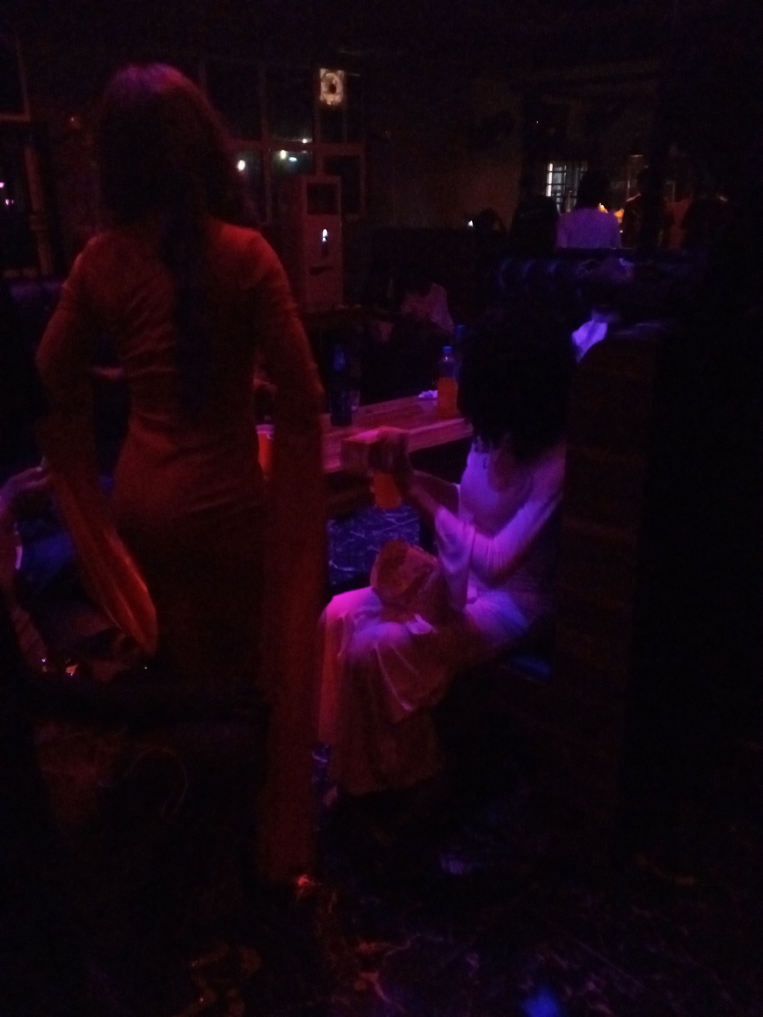 In a dimly lit indoor venue, possibly a bar or club in Lokoja, Nigeria, two women are visible. The woman on the left is standing and facing away from the camera, wearing a long red dress with wide sleeves. She appears to be holding an object in her left hand. To her right, a second woman is seated, facing left and slightly away from the camera. She is wearing a light-colored, possibly white or pink, garment and her right arm is extended as she holds a bottle or glass. The environment is dark with some ambient lighting creating purple and red hues, suggesting a nightlife setting. Tables and other patrons are partially visible in the background, contributing to a social atmosphere.