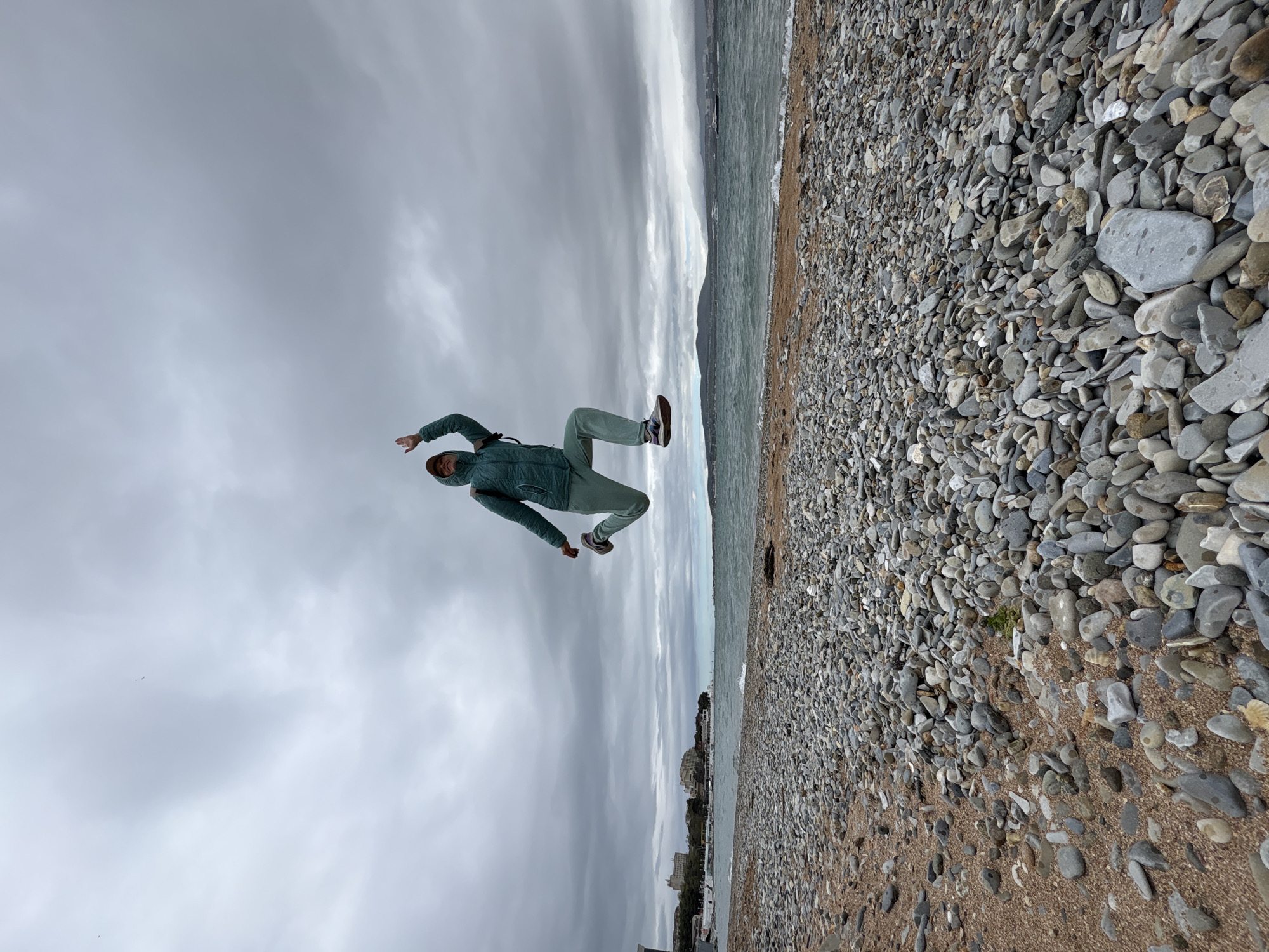 The image shows a person in mid-air, wearing a light green tracksuit and brown shoes, seemingly jumping on a beach. The location appears to be a beach covered with grey pebbles, with a body of water adjacent, possibly the sea. The sky above is overcast with grey clouds. In the distance, there are buildings visible, suggestive of a city. The buildings suggest a coastal city of Unknown, Unknown.