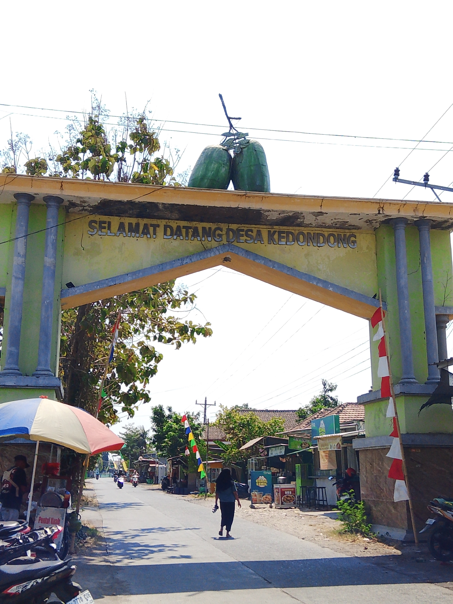 The image shows an archway in Kedondong, Indonesia, welcoming visitors to the village. The arch is painted light green and yellow, with decorative pillars on the sides. The words "SELAMAT DATANG DESA KEDONDONG" are written in silver letters on the upper part of the arch. Atop the arch, two large, green, elongated objects resembling melons are placed as decoration, along with some greenery. Indonesian flags, red and white, are attached to the right side of the arch.
In the foreground, a street scene unfolds. On the left, a stall with a red, white, and blue umbrella is set up, with motorcycles parked nearby. A person wearing a red shirt is visible behind the stall. A street stretches into the distance, lined with small shops and houses. Several motorcycles are on the street, some in motion. On the right side of the street, more shops with colorful signs are visible. A person in dark clothing is walking away from the camera down the street. Trees and power lines are present throughout the scene. The sky is bright and clear.
