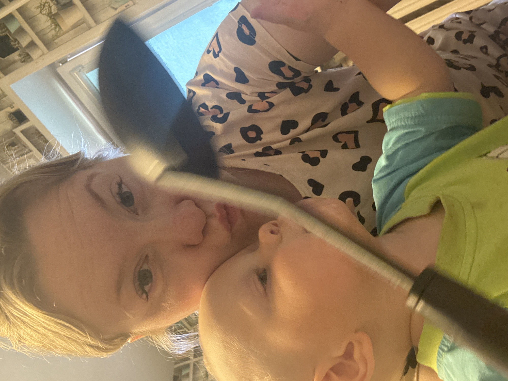 A woman with blonde hair and green eyes holds a young child. The woman wears a leopard-print shirt, and the child wears a green and blue shirt. A black object is in the foreground, partially obscuring the view. The background includes a window. The location is implied to be Chaikovsky, Russia.