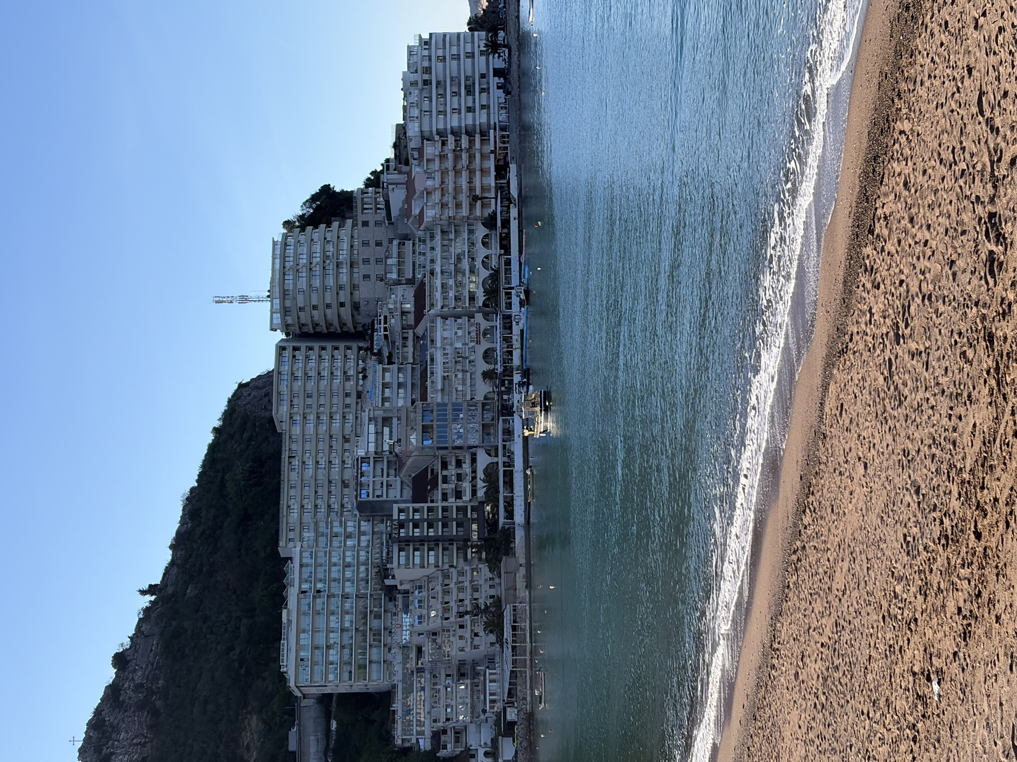 The image shows a coastal scene in Rafailovići, Montenegro. A densely packed cluster of high-rise buildings clings to a hillside beside a beach. The buildings are predominantly white with numerous windows and balconies. The dark green, wooded hillside rises sharply behind them.
Adjacent to the buildings is the calm, turquoise water of the Adriatic Sea with small waves breaking along the sandy beach. The beach is sandy, covering the foreground. The sky above is a clear blue. The overall scene suggests a popular tourist location.