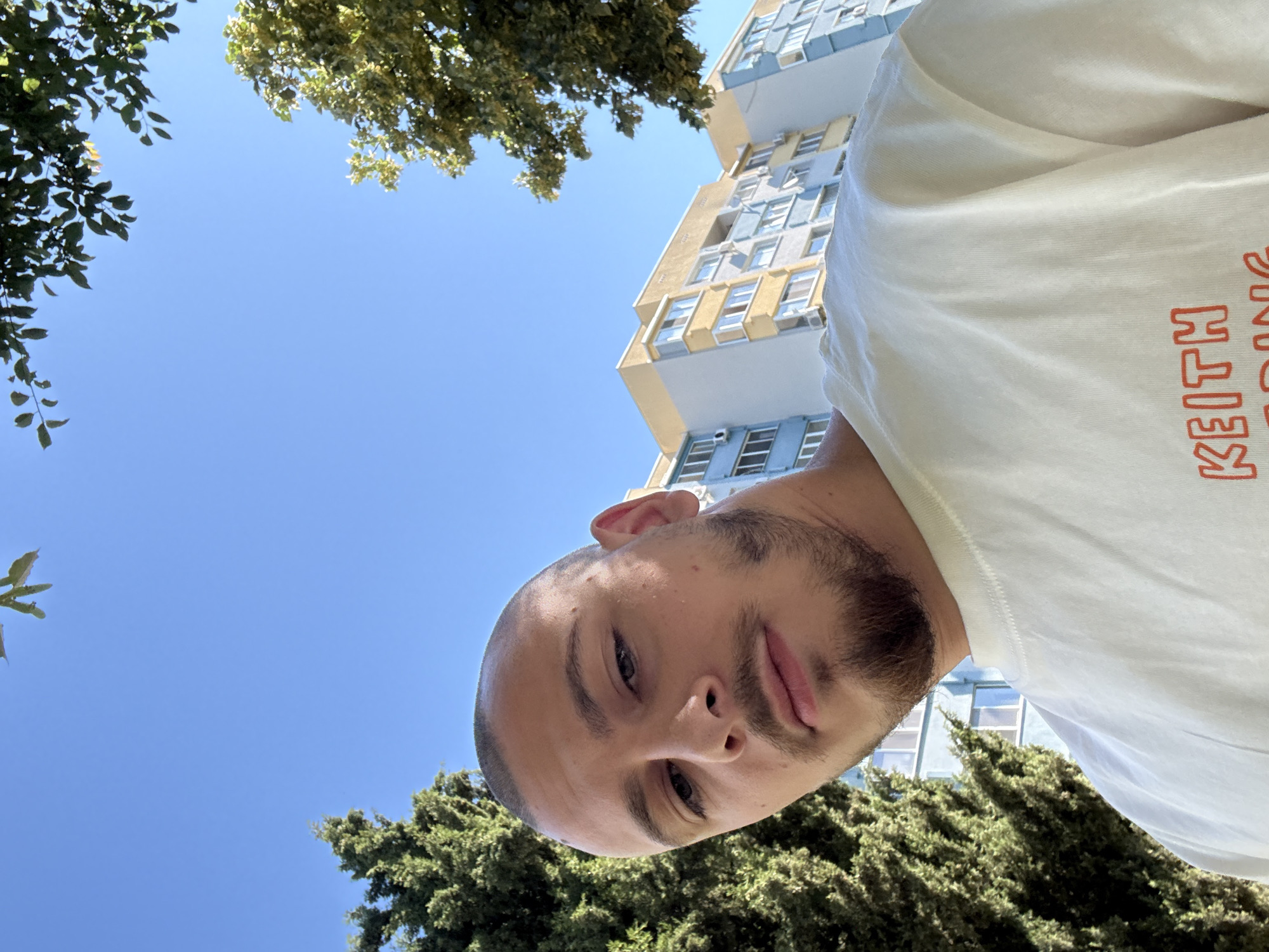 An individual with a shaved head and stubble is captured in a low-angle, outdoor selfie. The person is wearing a light-colored t-shirt with orange lettering that reads "KEITH HARING" on the chest. Behind the individual, a multi-story apartment building with light blue and yellow accents is visible, alongside lush green trees. The sky is a clear, bright blue, suggesting a sunny day. The scene is set in the city of Stara Zagora, Bulgaria, likely in a residential area given the apartment building and trees.