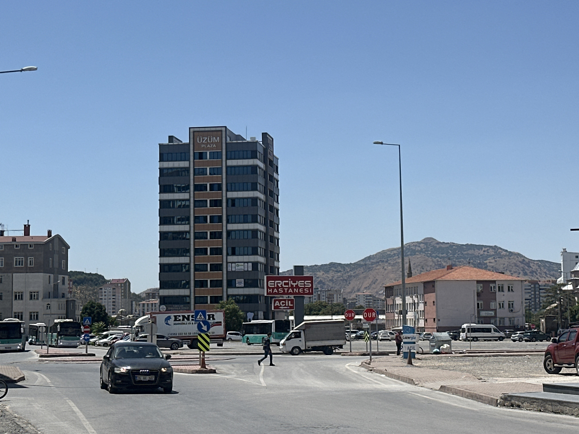 In Kayseri, Turkey, a street scene unfolds under a clear blue sky. A tall, modern building, labeled "UZUM PLAZA," dominates the mid-ground, its grey facade contrasted with bands of brown and numerous windows. Across the street, a sign for "ERCIYES HASTANESİ" (Erciyes Hospital) is visible, with smaller text below reading "ACİL" (Emergency). A black Audi sedan is driving through an intersection in the foreground, its headlights on. To the left, two buses are stopped at what appears to be a bus station. A lone pedestrian is crossing a crosswalk between the buses and the hospital sign. In the background, residential buildings and a large, arid mountain range suggest the city's geographical setting. Traffic signs, including "DUR" (Stop) signs, are present. The environment appears to be an urban commercial and residential area.
