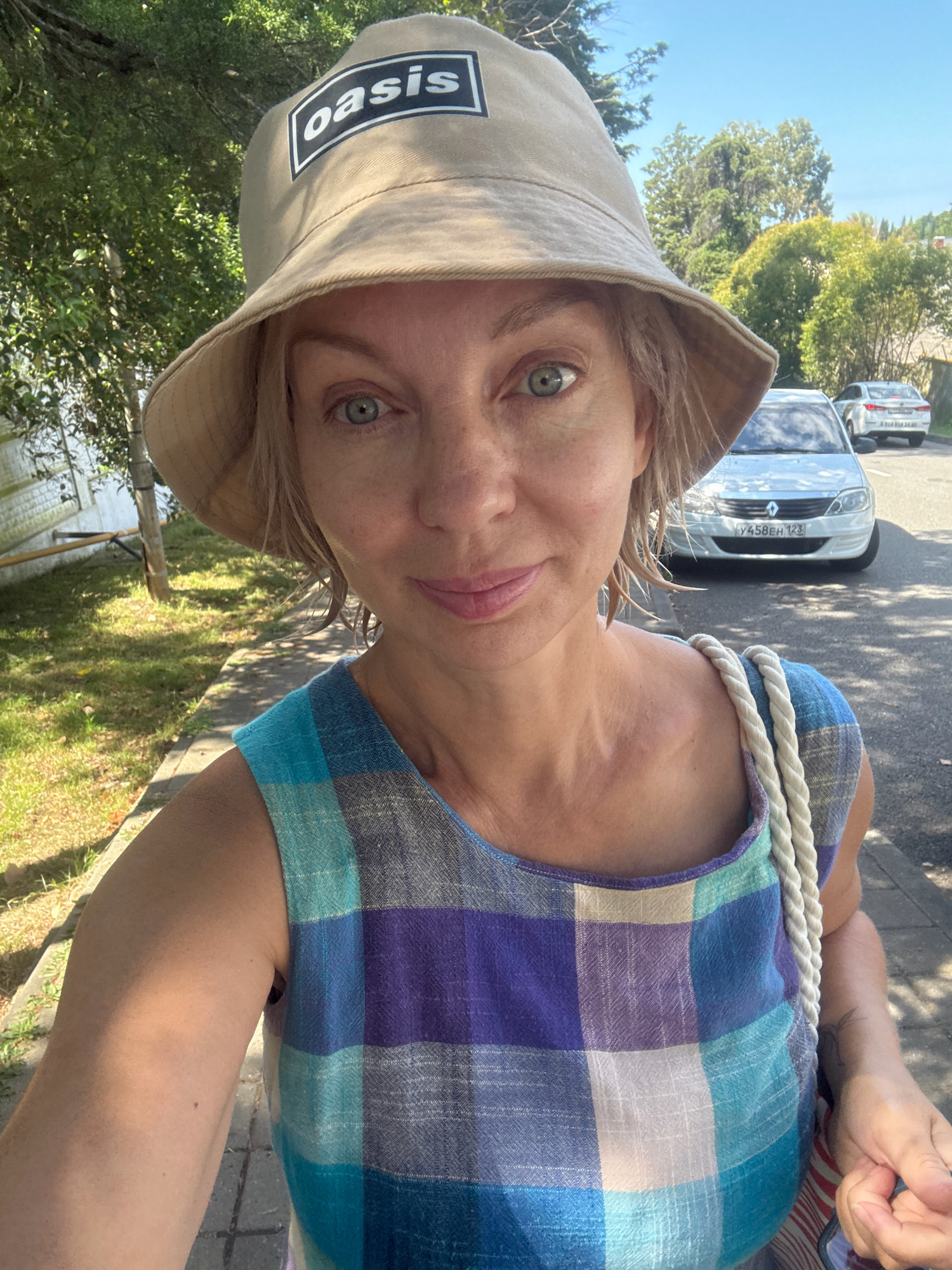 The image is a selfie taken by a woman outdoors, likely on a sunny day in Sochi, Russia. She is wearing a light-colored bucket hat with an "oasis" logo and a sleeveless dress with a plaid pattern in shades of blue, purple, and white. A thick white rope strap is visible over her shoulder, suggesting she is carrying a bag. In the background, a white car with a Russian license plate is parked on the street, and there are trees and greenery lining the sidewalk. The woman's expression is neutral to pleasant, and she is looking directly at the camera.