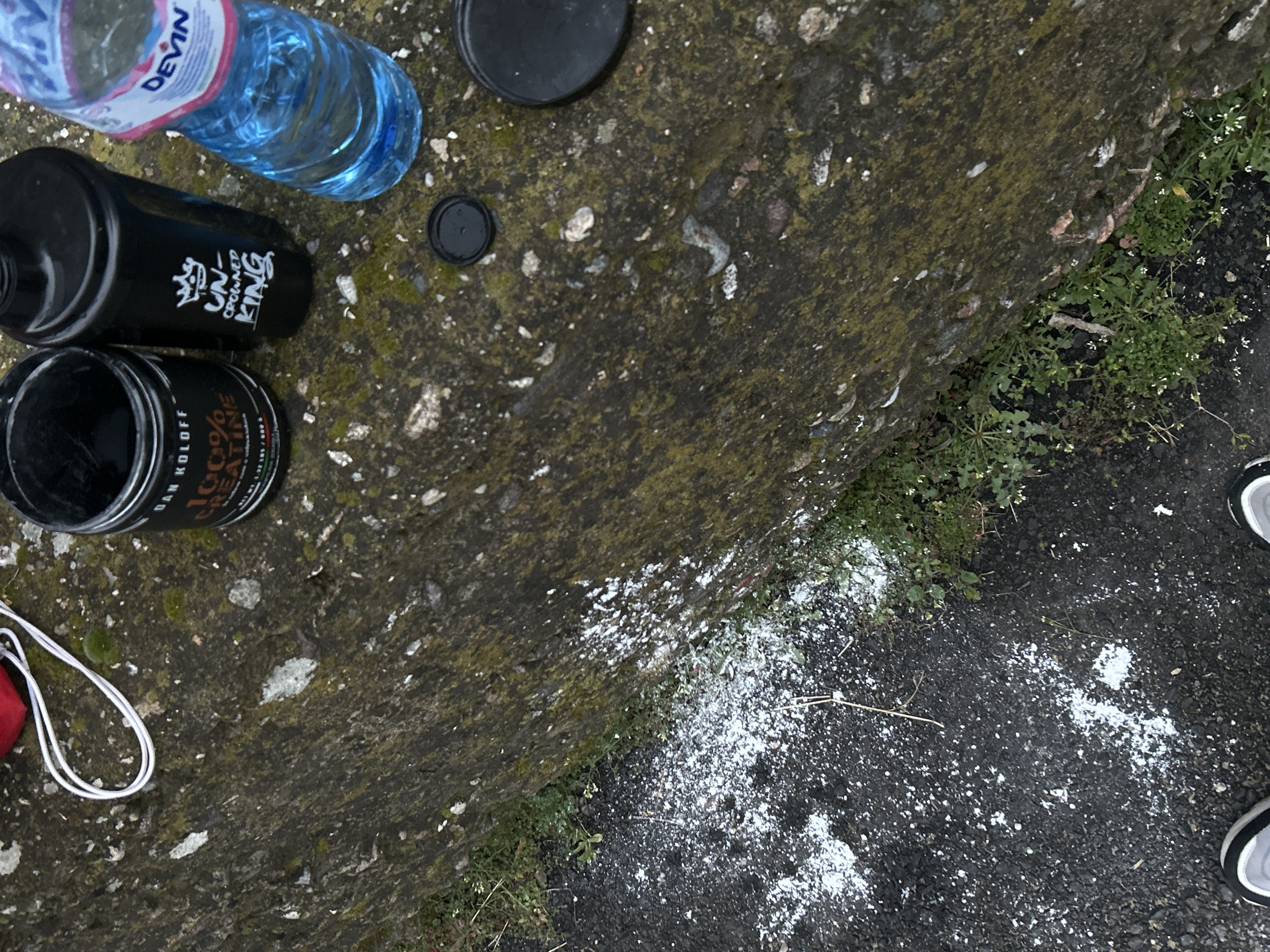 The image shows an outdoor scene on what appears to be a weathered concrete surface, possibly a sidewalk or the edge of a road. The setting is in Zlatograd, Bulgaria. Several objects are present: a plastic water bottle labeled "Devin", a black cylindrical bottle marked "Uncrowned King", a container of "Dan Koloff 100% Creatine", and two black bottle caps. A white powder substance is scattered on the adjacent dark asphalt surface, along with some sparse vegetation growing between the concrete and asphalt. Part of a white drawstring bag is visible near the creatine container. Two sneakers are visible on the right edge of the image. There are no visible human figures, only feet wearing the sneakers. The environment shows signs of neglect and debris.