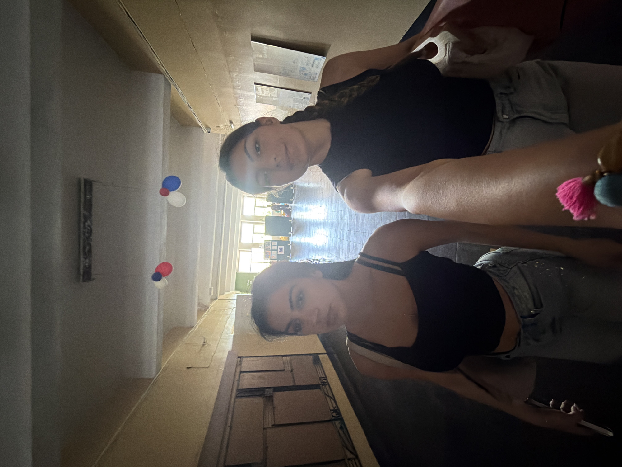Two young women are featured in what appears to be a selfie, captured indoors in a long hallway or large room, likely in Burgas, Bulgaria. The original image is rotated.
The woman in the foreground wears a black strappy top and light-colored jeans or shorts, with dark hair cascading over her shoulder. She exhibits a subtle smile. A keychain with pink and teal tassels hangs from her wrist or is held in her hand, which grasps a white object, possibly a smartphone. The second woman, positioned behind and slightly to the left, also has dark hair and is dressed in a black top and light-colored jeans. She looks directly at the camera with a neutral expression.
The setting is a well-lit indoor corridor with high ceilings, light-colored walls, and a light-toned floor, possibly wood or linoleum. Columns or pillars are visible along the hallway. Bright natural light illuminates the space from windows at the far end, indicating it is daytime. On the left side of the frame, two clusters of balloons – each comprising red, white, and blue balloons – float in the air. Given the location context of Burgas, Bulgaria, these colors strongly suggest a connection to Bulgarian national symbols or a patriotic celebration. Framed artwork is visible on the walls, and some furniture or display units are present near the distant windows. The environment suggests a public or institutional building, such as a school or community center, possibly decorated for an event. No visible text is discernible in the image.