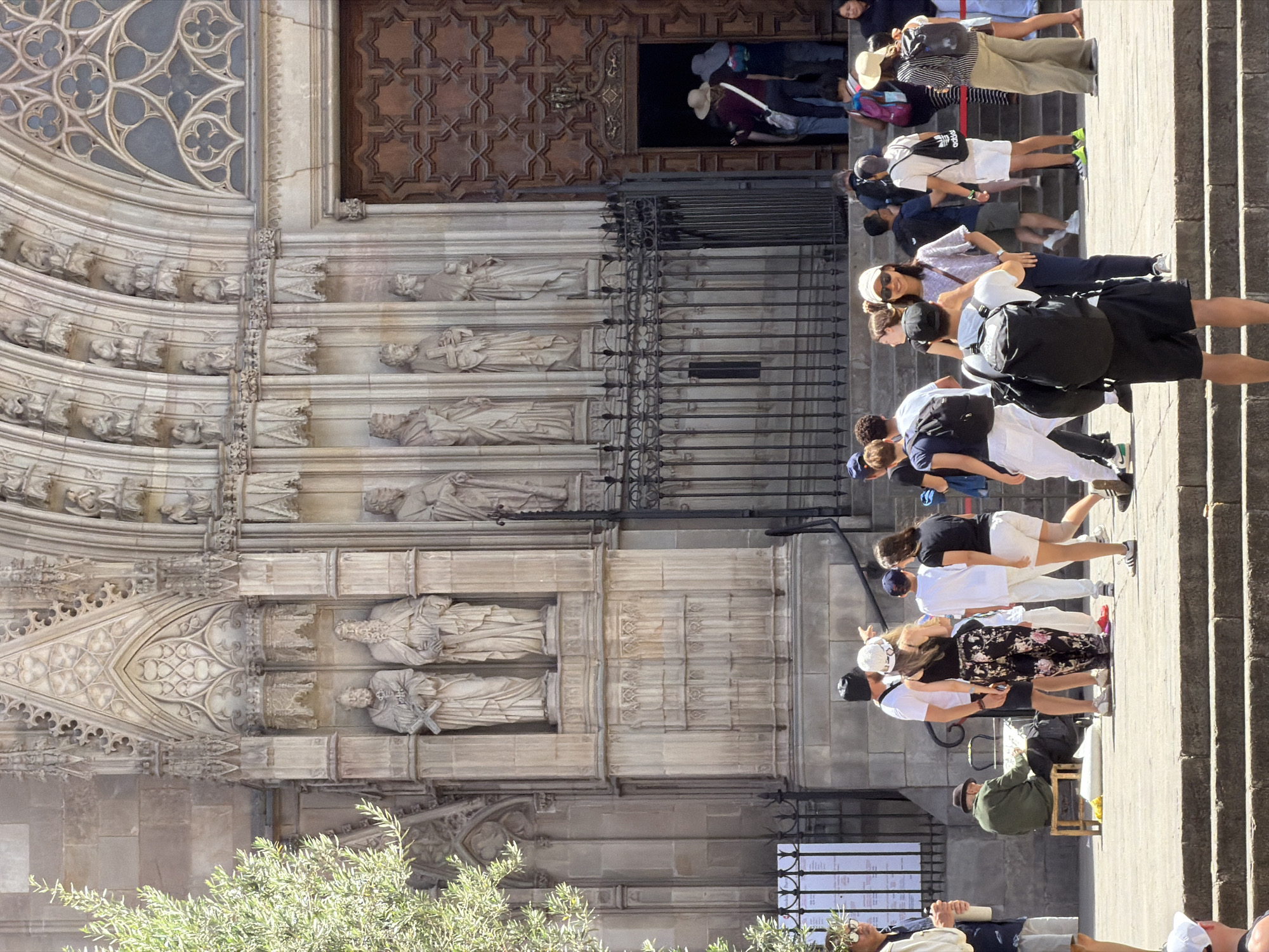 The image depicts a crowd of tourists gathered on the steps and plaza in front of the Barcelona Cathedral, located in the Gothic Quarter of Barcelona, Spain. The cathedral's facade, characterized by intricate Gothic architecture, ornate sculptures of figures in niches, and a large, decorative wooden door, is prominently featured. The scene is set on a sunny day, with shadows cast by the architecture and people.
Several individuals are visible in the foreground and mid-ground. Some are standing, others are walking, and a few are seated. A man in a green jacket is seated on a small stool on the plaza, appearing to be an artist or performer. Tourists are dressed in casual summer attire, with many carrying backpacks and wearing hats for sun protection. Some people are looking at the cathedral, while others are interacting with each other.
The environment includes stone steps leading up to the plaza, a black metal fence partially enclosing an area, and a large, ornately carved wooden door that is slightly ajar, with people entering. The architectural details of the cathedral are sharp and well-defined, suggesting a clear and bright day. The crowd appears to be diverse, typical of a popular tourist attraction.