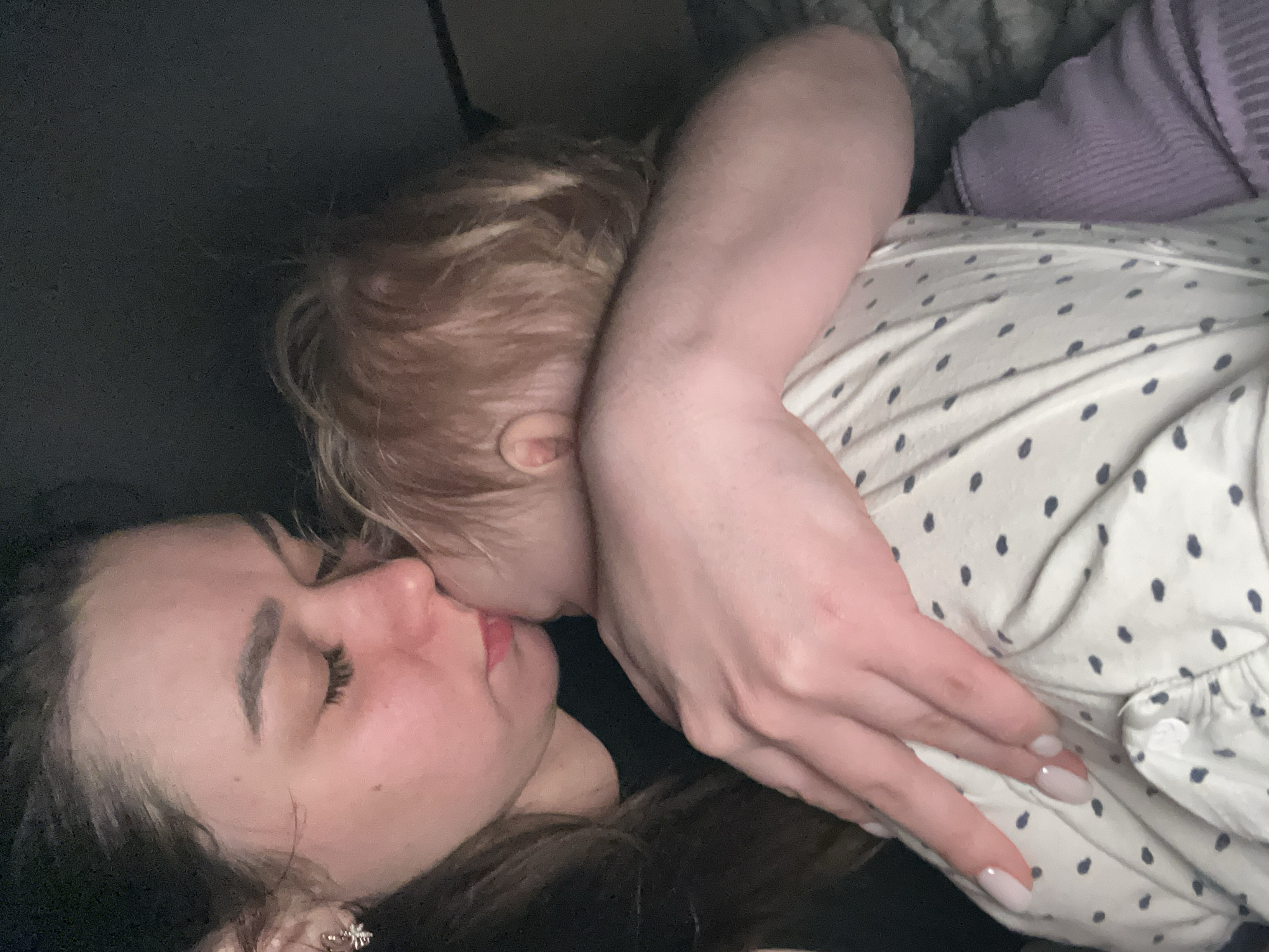 This image captures a close-up, intimate moment between an adult female and a young child, likely indoors in Chaikovsky, Russia. The perspective is angled downwards, showing the two subjects nestled together, implying a moment of rest or affection.
The adult, a female with dark hair, lies on her side with her eyes closed, suggesting sleep or deep relaxation. Her lips are slightly parted. She has prominent eyebrows and visible eyelashes. A small, sparkling stud earring is visible in her right ear. Her left hand, with light-colored, oval-shaped manicured nails, rests gently on the child's torso.
The child, with light-colored (possibly blonde or light brown) hair, is positioned very close to the adult's face, appearing to be kissing or nuzzling the adult's mouth or chin. Only the back of the child's head, an ear, and part of their arm and torso are clearly visible.
The adult is wearing dark clothing. The child is dressed in a light-colored garment, possibly a sleepsuit or top, adorned with a pattern of small, dark blue, teardrop-shaped dots. A section of soft, purple, ribbed fabric, possibly a blanket or part of another garment, is visible in the background near the child.
The overall scene conveys a strong sense of tenderness, comfort, and maternal affection. The dim lighting suggests it is likely evening or nighttime, contributing to the serene and intimate atmosphere. There is no visible text in the image.