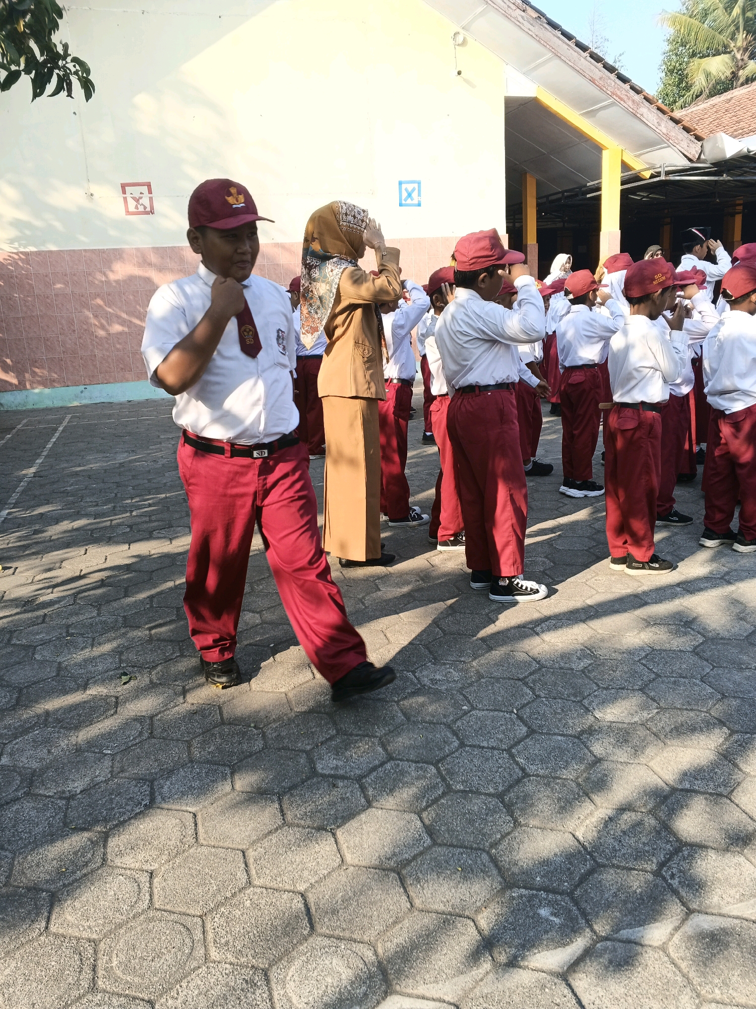 In Glagah, Indonesia, a group of schoolchildren in uniform are standing on a paved outdoor area, possibly a schoolyard. The children are wearing white shirts, maroon ties, and maroon pants or skirts, and many have maroon caps. A few adults, likely teachers, are also present. One adult woman in a tan hijab and brown uniform stands in the foreground, her hands raised. The schoolchildren appear to be engaged in an activity, with some holding their hands up to their foreheads, suggesting a pose or salute. The environment is sunny with dappled shadows on the hexagonal paving stones. A building with a yellow facade and a red-tiled roof is visible in the background.