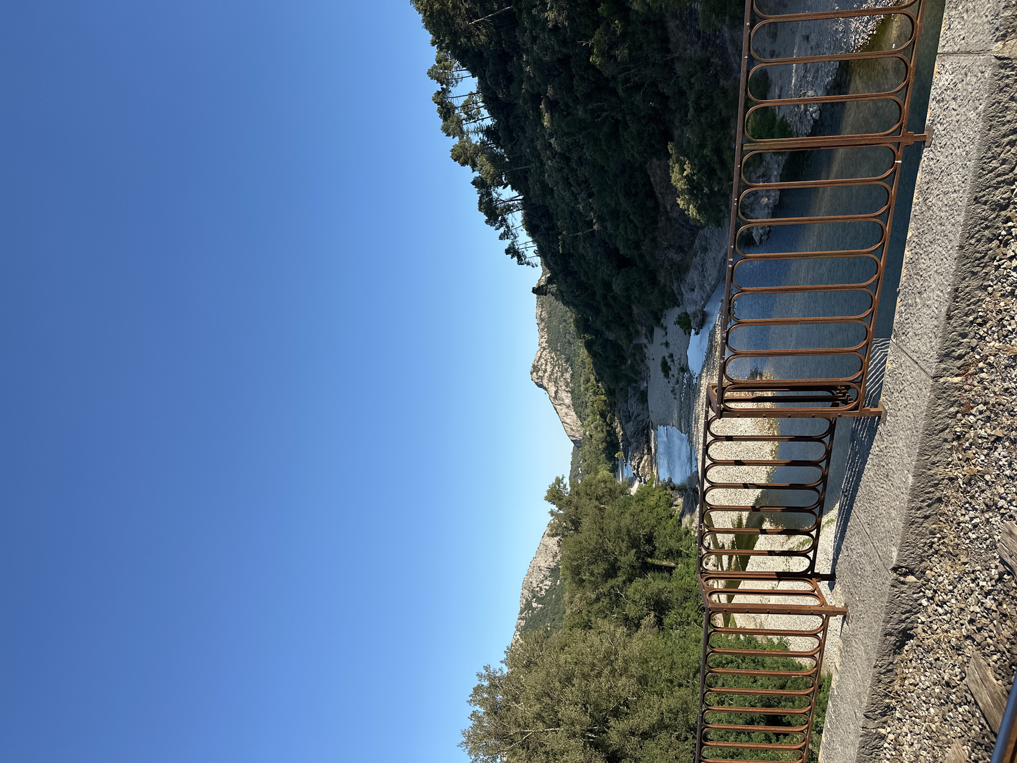 This image, captured from a bridge in Anduze, France, depicts a picturesque view of the Gardon River flowing through a valley. The perspective is from the bridge's railing, which is made of rusted metal with an ornate, looped design, and sits atop a concrete structure covered in gravel. Below the railing, the river winds through a rocky, pebble-strewn bed, with clear, reflective water. Lush green trees and vegetation cover the slopes of the surrounding hills, with rugged, rocky mountain peaks visible in the distance under a clear blue sky. The scene suggests a natural, serene landscape, likely in the afternoon given the quality of light and shadows. No people are visible in the image.