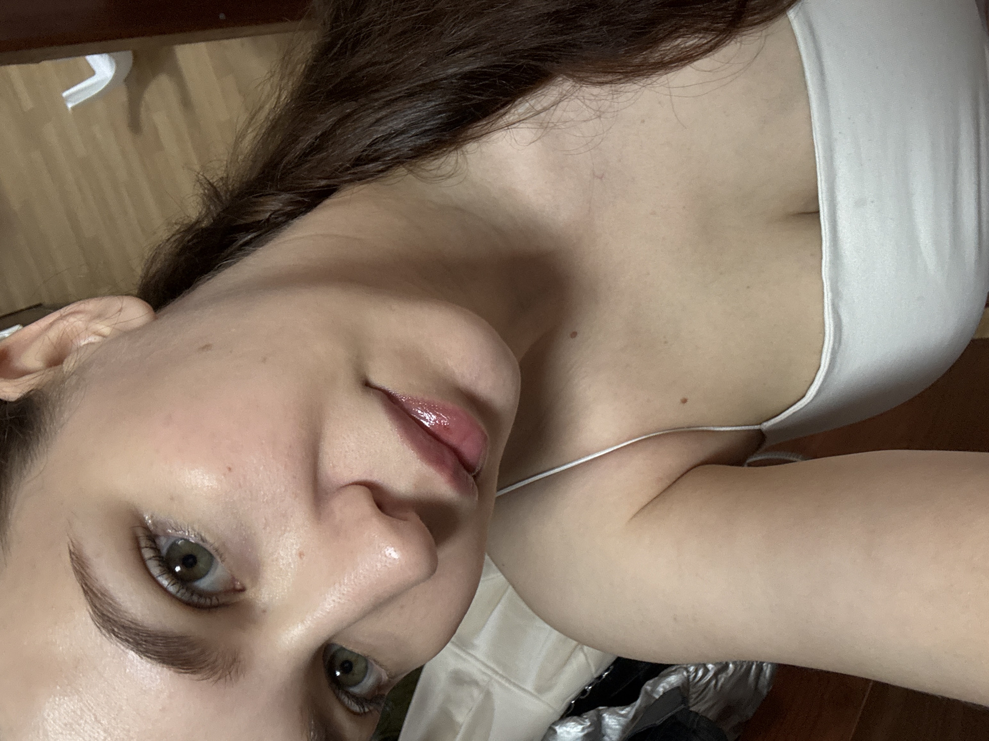 An intimate, rotated portrait features a young woman indoors in Chaikovsky, Russia. Her head occupies the left side of the frame, with her body extending to the right. She has fair skin, prominent green/hazel eyes, and neatly shaped dark eyebrows. Her full lips are enhanced with a glossy finish. Her long, dark brown hair cascades around her head and shoulders. She wears a simple white top with thin straps.
The woman exhibits a subtle, gentle expression, gazing directly towards the camera. The setting appears to be a bedroom or living area with wooden floorboards visible in the background, along with what seems to be a dark piece of furniture and some crumpled white material (possibly paper or fabric) near her head. Another crumpled, metallic-looking object is partially visible near her right arm. The lighting is soft and diffused, highlighting her features without harsh shadows. There is no visible text in the image.