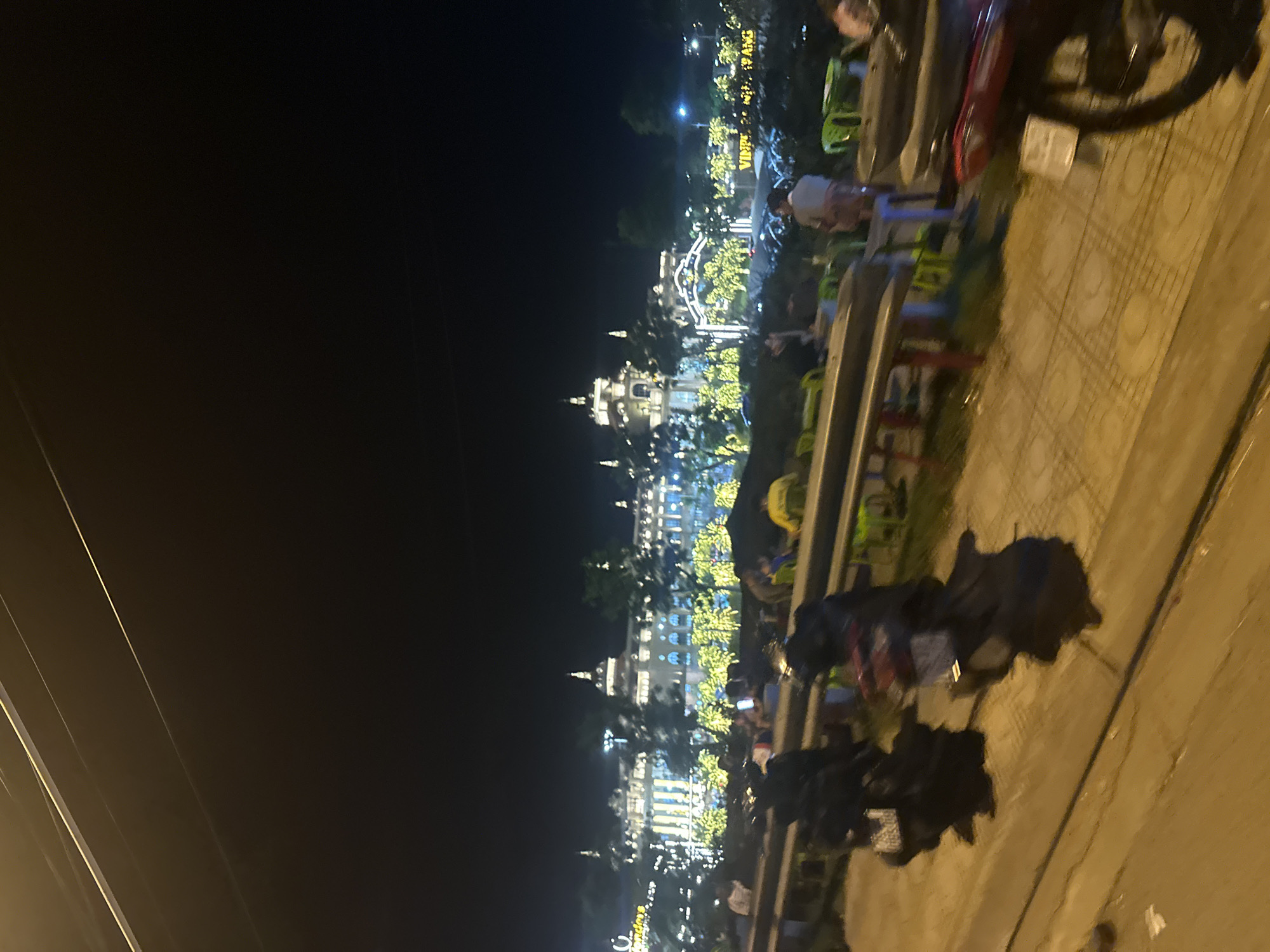 The image depicts a nighttime scene in Nha Trang Ward, Vietnam, likely along a waterfront promenade or street. In the background, a grand building, possibly a hotel or entertainment venue, is illuminated with numerous yellow lights, creating a festive atmosphere. The architecture features ornate details and multiple domes. Trees decorated with similar yellow lights are scattered in front of the building, enhancing the festive ambiance.
In the foreground, a row of green plastic chairs and tables are set up, suggesting an outdoor seating area for dining or socializing. Several people are seated at these tables, though their faces are indistinct due to the low light and motion blur. Motorbikes are parked along a pathway in front of the seating area, with some covered by dark tarps. One motorbike, partially visible on the right, is red and appears to be a scooter. The ground is paved with patterned tiles. The overall impression is of a vibrant evening scene in a popular urban area of Nha Trang.