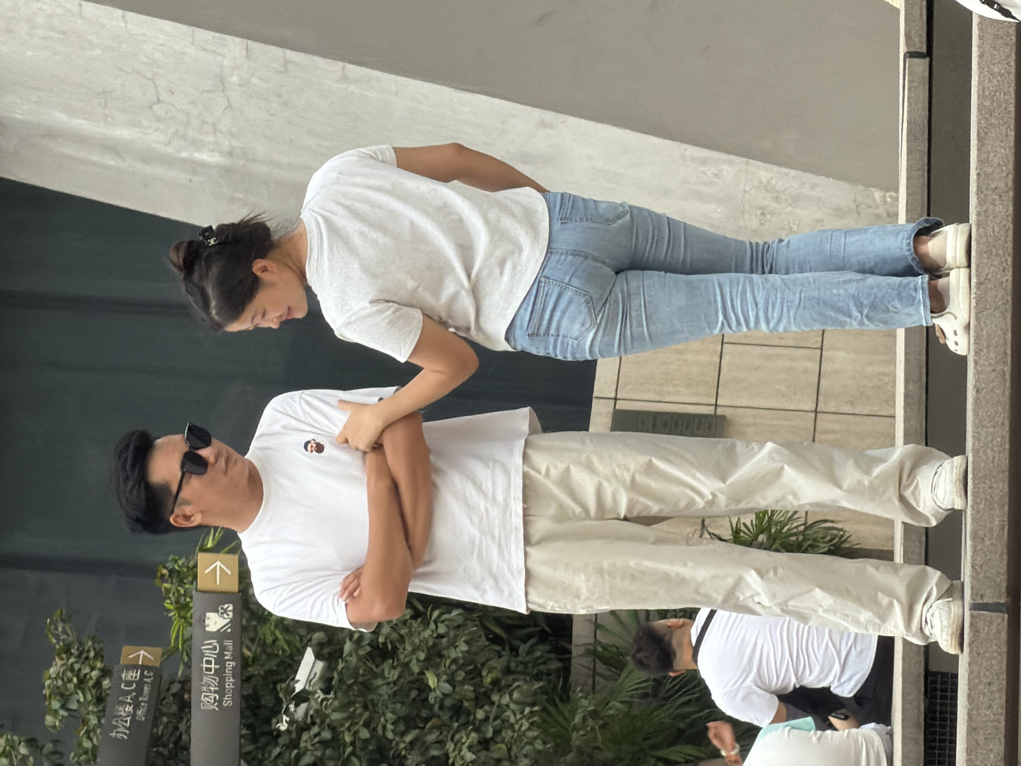 In Yuzhong District, China, a man and a woman are standing on a step. The man, wearing sunglasses, a white t-shirt with a cartoon graphic, and light-colored pants, has his arms crossed. The woman, with her hair tied up, is wearing a white t-shirt and blue jeans. She is reaching out to touch the man's arm. In the background, there are directional signs for "Office Tower A, C" and "Shopping Mall." Lush green foliage is visible to the left, and a textured concrete structure is on the right. Another person is seated in the lower left corner of the frame, facing away from the camera.