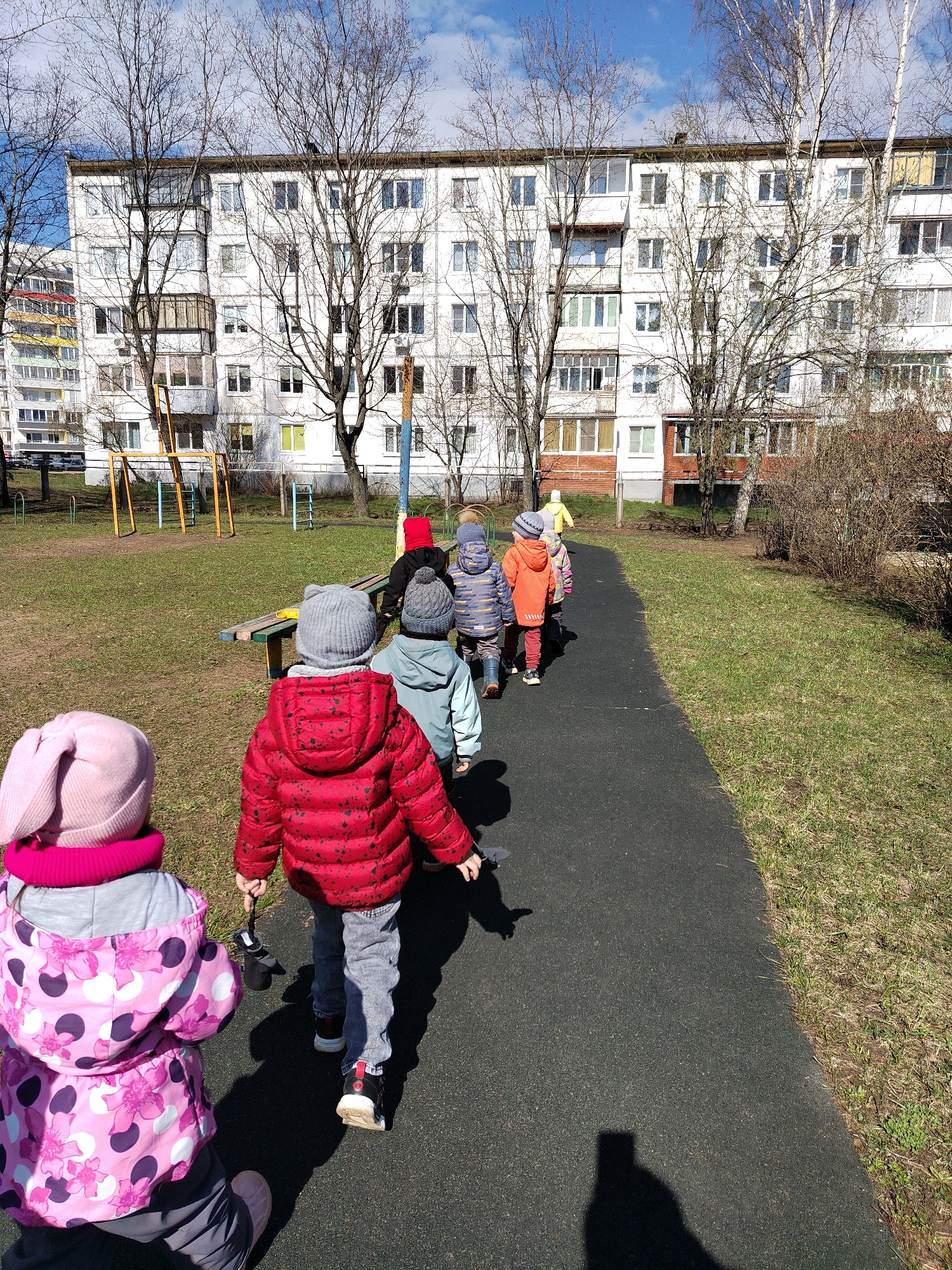 A group of young children are walking in a line on a paved path in an outdoor area. The children are wearing winter clothing, including jackets, hats, and pants. The location is Chaikovsky, Russia, as can be inferred from the architecture of the building in the background, which is a large, white apartment building with a repetitive window pattern. There are bare trees scattered throughout the scene, and a playground is visible to the left. The ground is a mixture of grass and dry leaves, suggesting it is early spring. The children appear to be on a walk or outing, likely from a daycare or preschool. One child is holding gloves in their hand.