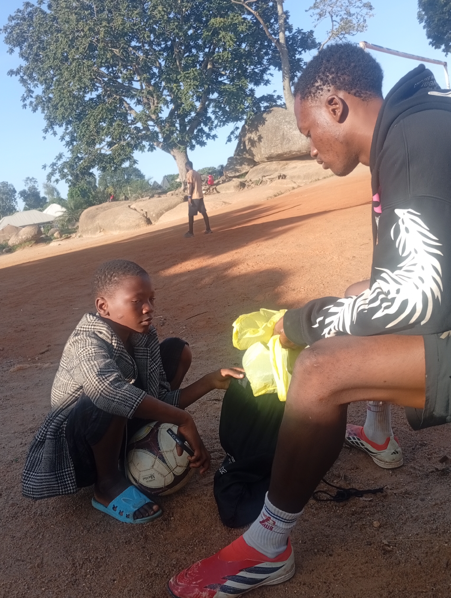Two young people are sitting on the ground in what appears to be a rural or outdoor area. The person on the left is a child wearing a patterned jacket and blue sandals. They are looking towards the person on the right and have a soccer ball in front of them, with a black pen or marker in their hand. The person on the right is an older youth or young adult wearing a black hoodie with white graphic designs on the sleeves and black shorts. They are holding a yellow plastic bag. The ground is covered in reddish-brown dirt and small stones. In the background, there are large rocks, trees, and some simple structures. The lighting suggests it is daytime, possibly late afternoon, given the shadows.