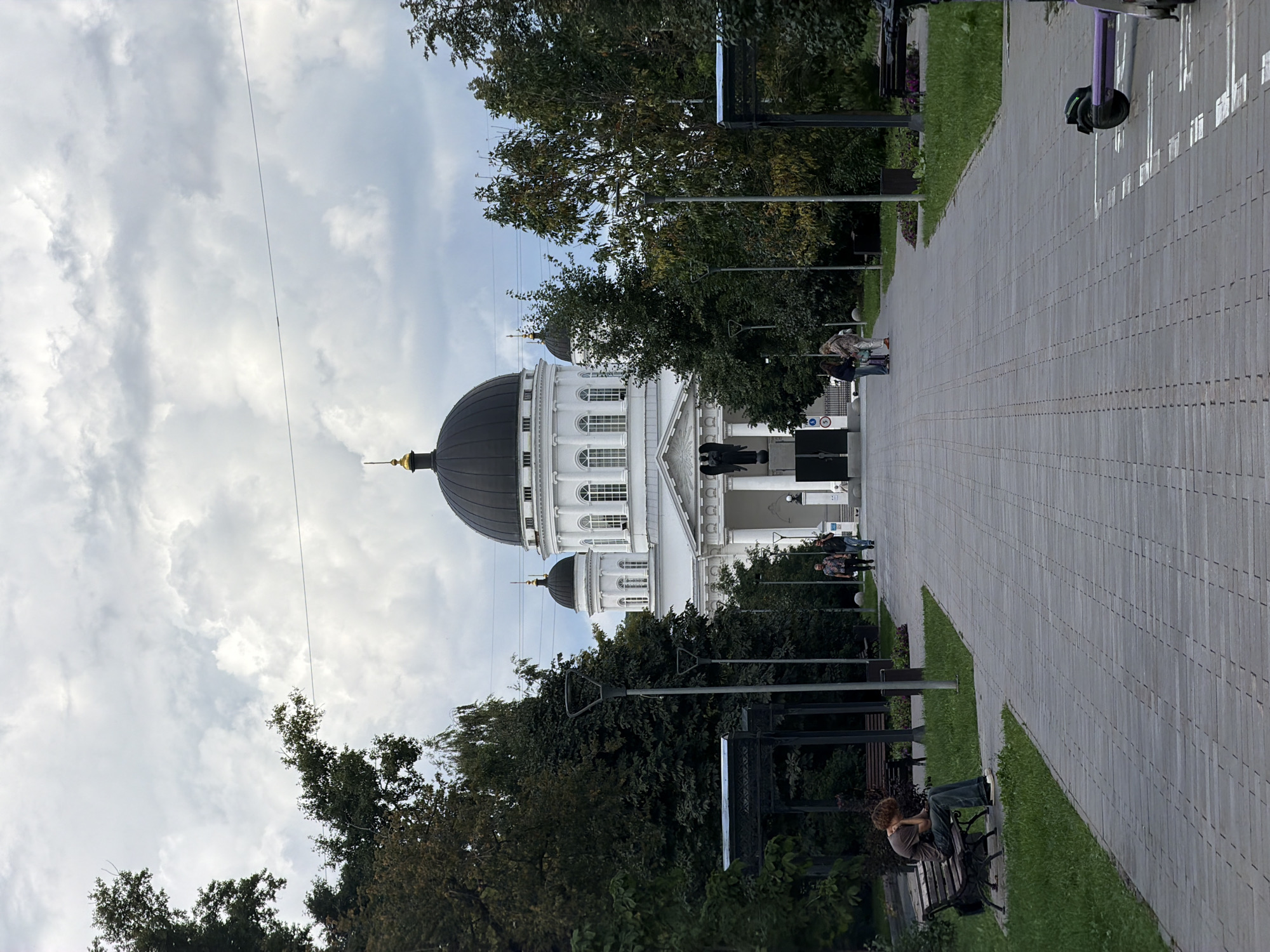 A wide, paved pathway leads towards a large, white domed building with classical architectural elements, such as columns and arched windows. The sky above is overcast with heavy clouds. On the left side of the pathway, a person sits on a bench facing away from the camera, appearing to be looking down at something in their lap. They are wearing a t-shirt and jeans. Further down the pathway, a group of three people are walking towards the building. On the right side of the pathway, several electric scooters are parked, marked by white lines on the pavement. The building is surrounded by trees, some of which have autumnal foliage. The overall atmosphere is somewhat somber due to the cloudy weather. The building is likely the Alexander Nevsky Cathedral in Nizhny Novgorod, Russia, given its distinctive architecture and prominent dome.