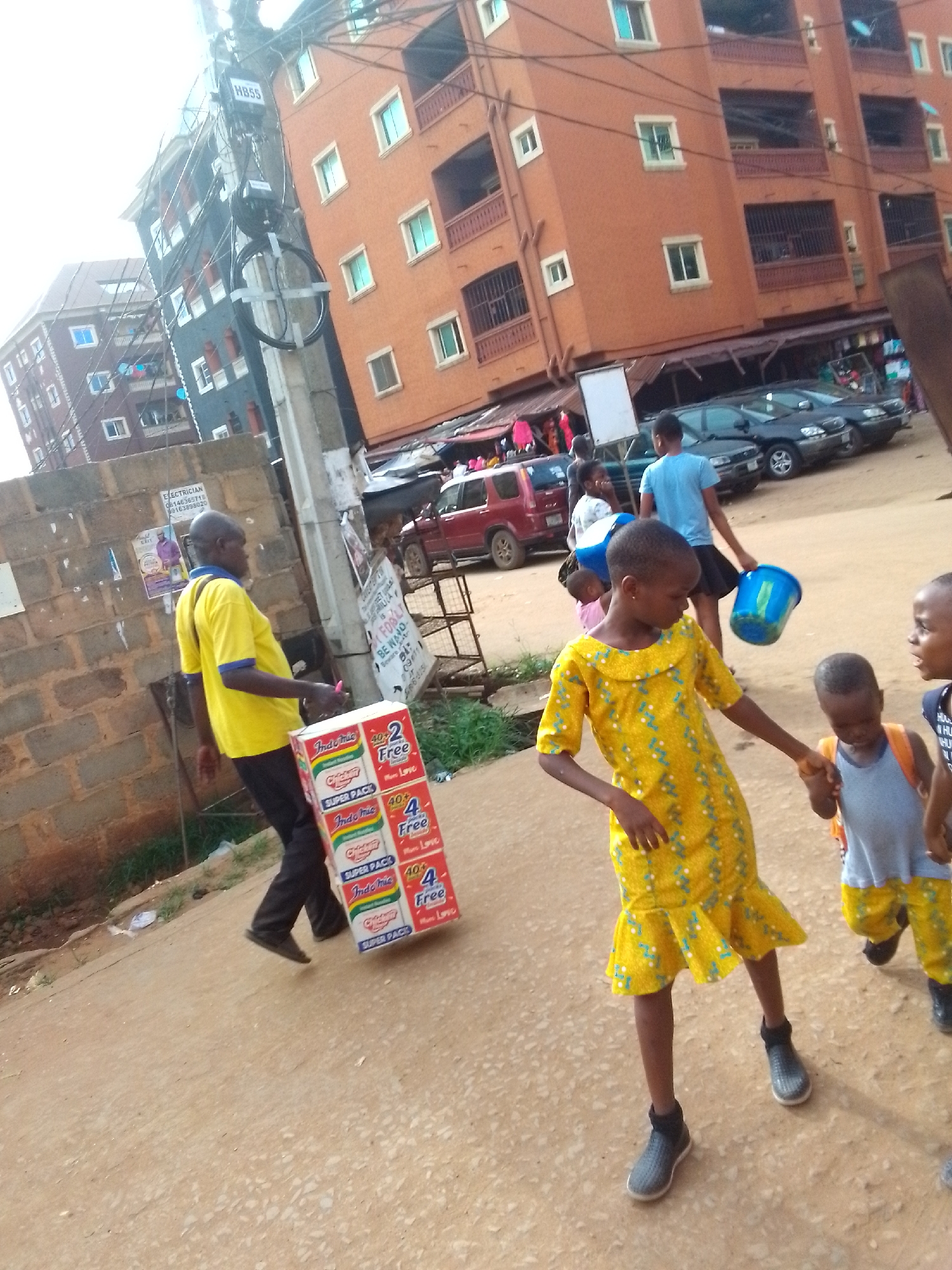 The image depicts a street scene in Onitsha, Nigeria. A man in a yellow shirt and dark pants carries a stack of Indomie noodle packages. Several children are present: one in a yellow dress and grey shoes holds the hand of another child wearing a blue tank top and yellow patterned shorts. Another person carries a blue bucket. Several cars are parked in the background and buildings loom in the distance. Power lines and a utility pole are visible near the buildings. Posters are adhered to the brick wall.