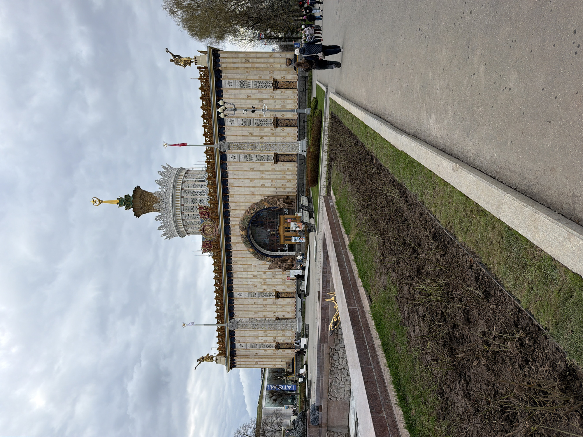 This image captures a grand, ornate building, likely a pavilion at an exhibition center in Moscow, under an overcast sky. The architecture is characterized by its Stalinist Empire style, featuring a multi-story structure with intricate detailing, decorative columns, and golden accents. A prominent central dome is topped with a large golden star and what appears to be a hammer and sickle emblem. Several gilded statues, including figures holding wreaths or tools, adorn the building's roofline. A small red flag is visible flying from a flagpole near the dome.
A paved pathway runs alongside the building, where several people are seen walking or standing casually, suggesting a public park or exhibition ground setting. Between the pathway and the building is a long, narrow garden bed with sparse vegetation, primarily bare soil and some green grass, indicative of early spring or late autumn. Trees are visible in the distance beyond the pathway.
Near the building's main arched entrance, a blue sign with white text that reads "ATOM!" is visible. The scene portrays a typical daytime atmosphere in a cultural or exhibition area, with the overcast weather providing diffused lighting. The architectural style, grandeur, and symbolic elements strongly point to a location such as VDNH in Moscow.